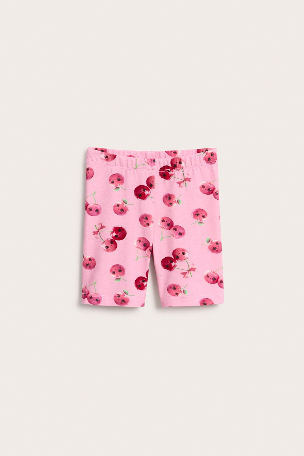 Framifrån: rosa barncykelbyxor med resår i midjan och körsbärsmönster, några med ansikten.