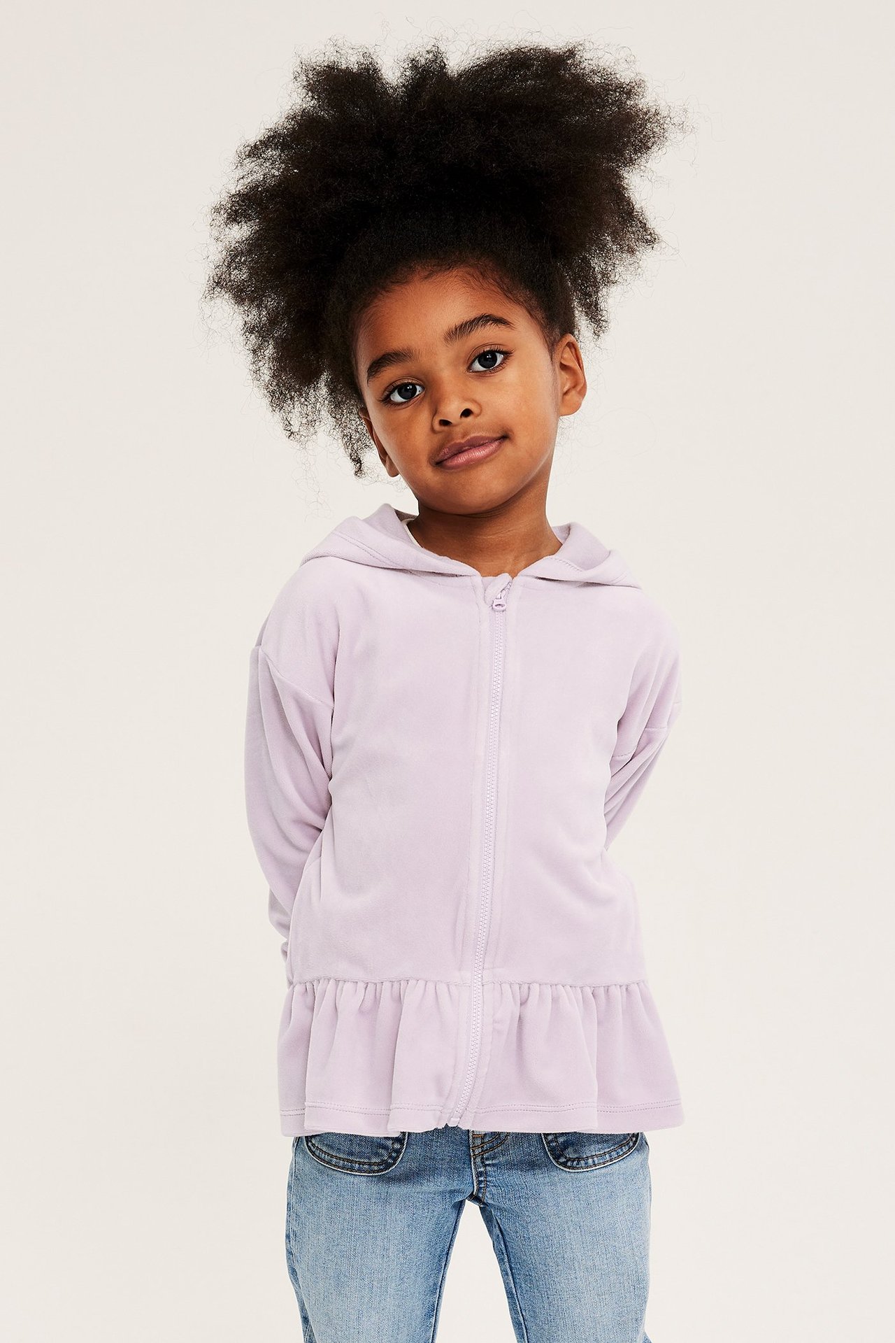 Forfra, et barn i en lys lilla velourhettegenser med glidelås, peplumkant og blå jeans.