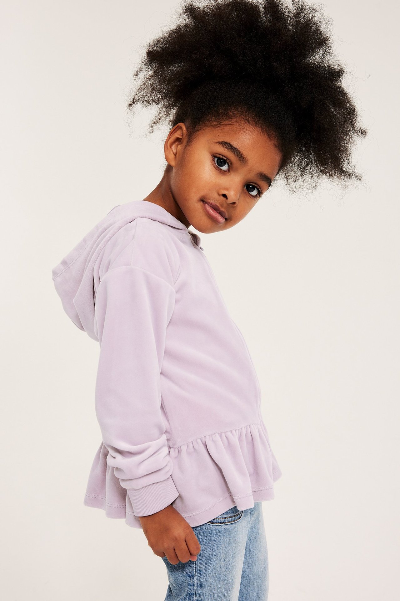 Sidovy av ett barn i ljuslila velourhoodie med peplumfåll och blå jeans.