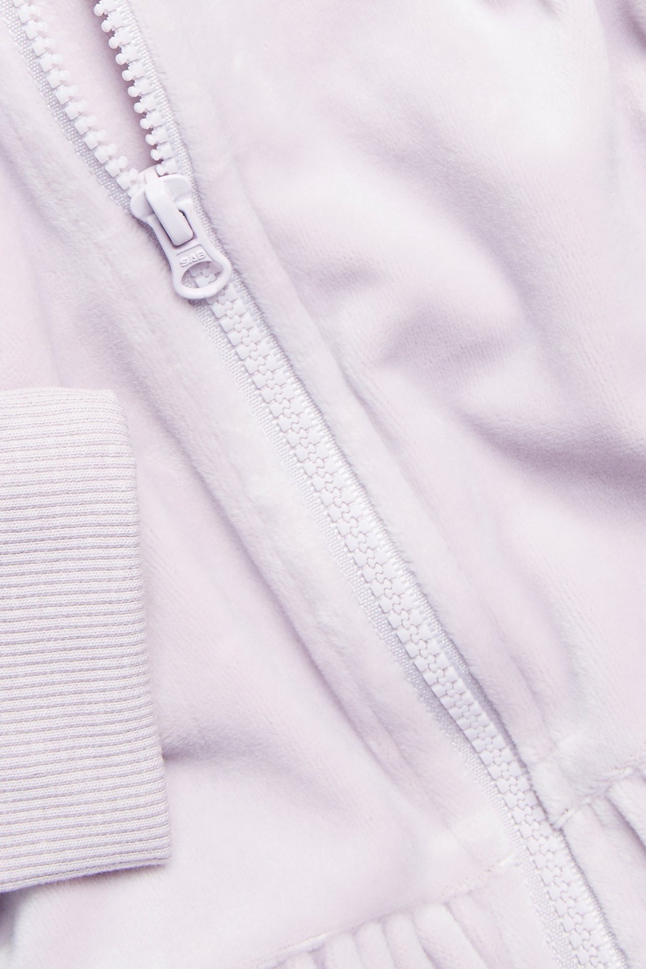 Zbliżenie jasnoliliowej welurowej bluzy z kapturem, z białym zamkiem, prążkowanym mankietem i subtelną falbanką.