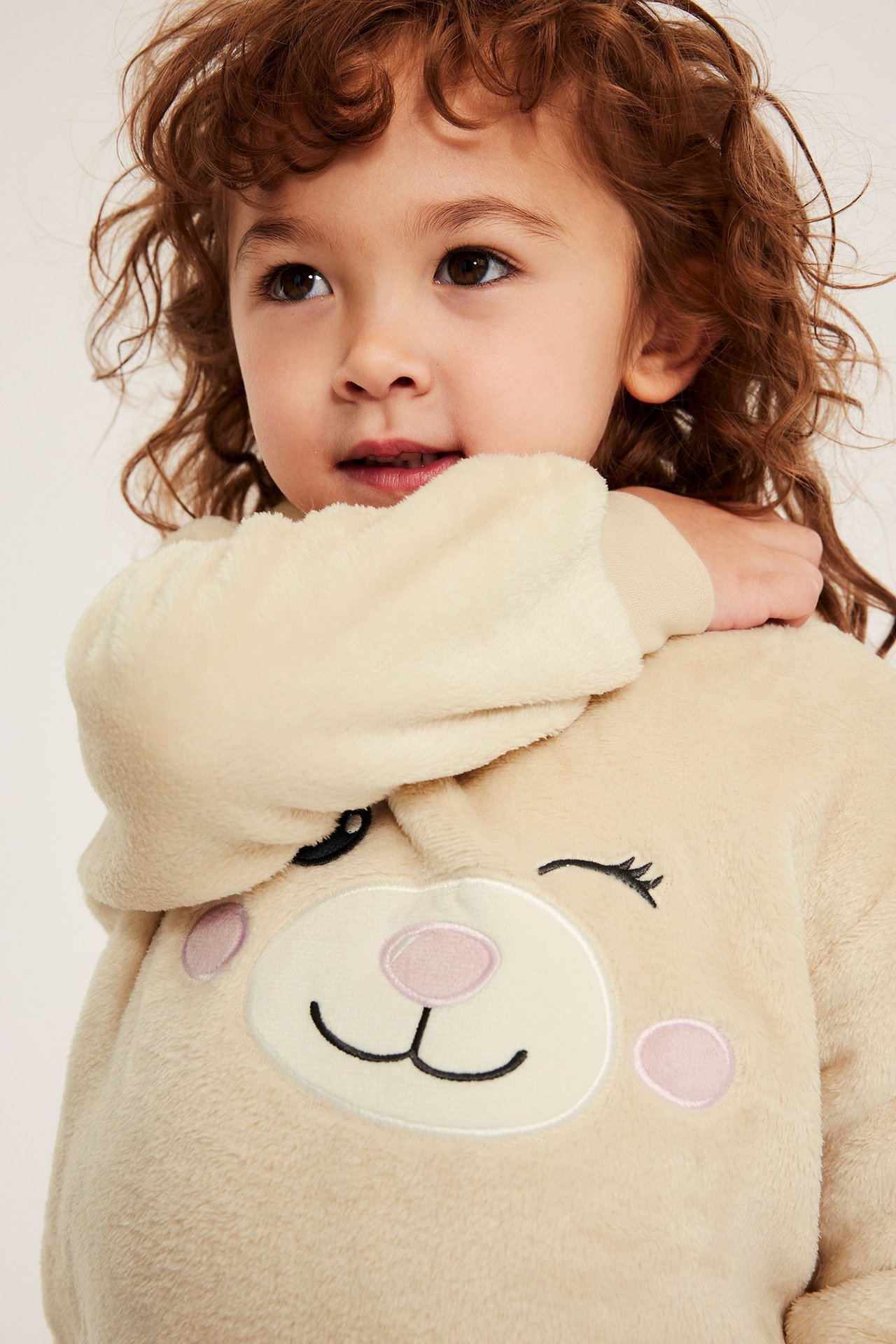 Lähikuva lapsesta vaaleassa beigessä pörröisessä hupparissa, jossa on silmää iskevä kasvoprintti edessä.