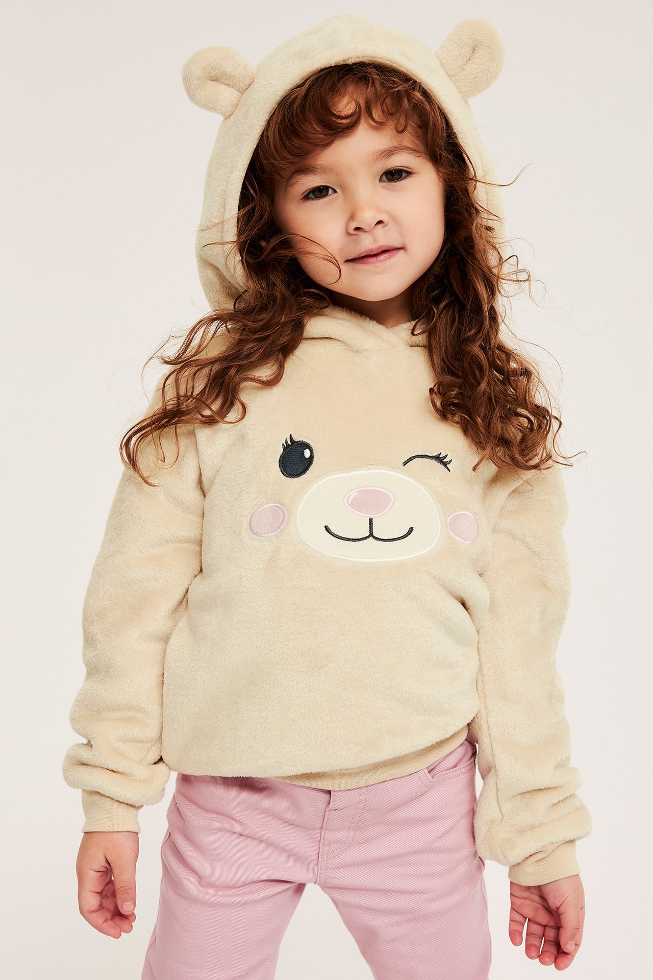 Frontvy, barn i fluffig ljusbeige hoodie med björnöron, blinkande björntryck och rosa byxor.
