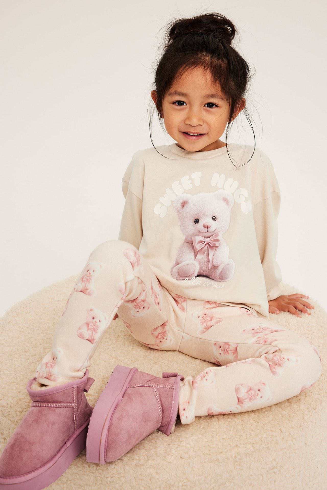 Barnkvinna i långärmad beige tröja med tryck av nallebjörn, rosa leggings och lila skor.