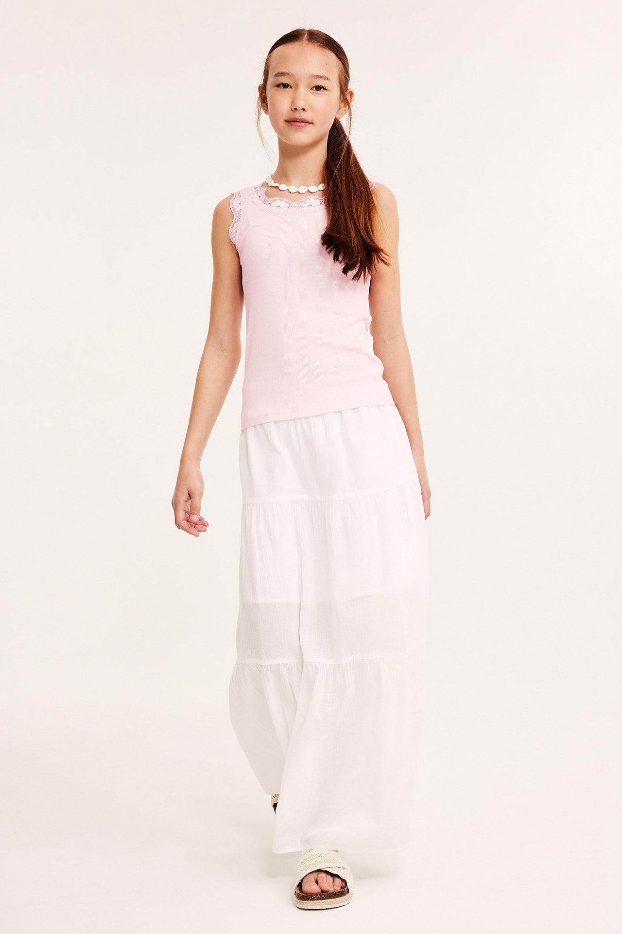 Framifrån: Barn i rosa ribbat linne med spets, vit volangkjol, vita sandaler och halsband.