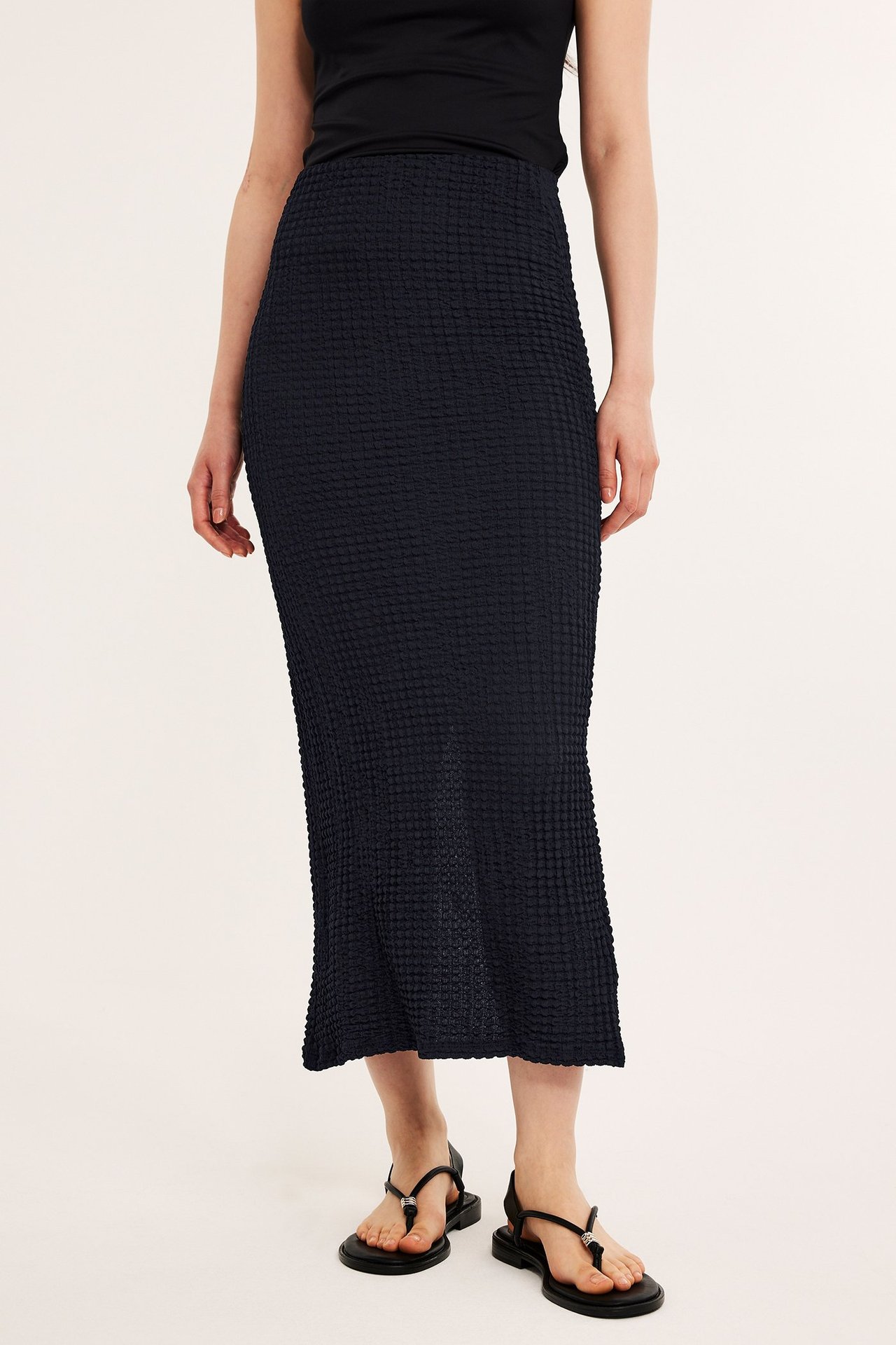 Frontvisning av en kvinne i langt svart strukturert skjørt, svart topp og svarte sandaler.