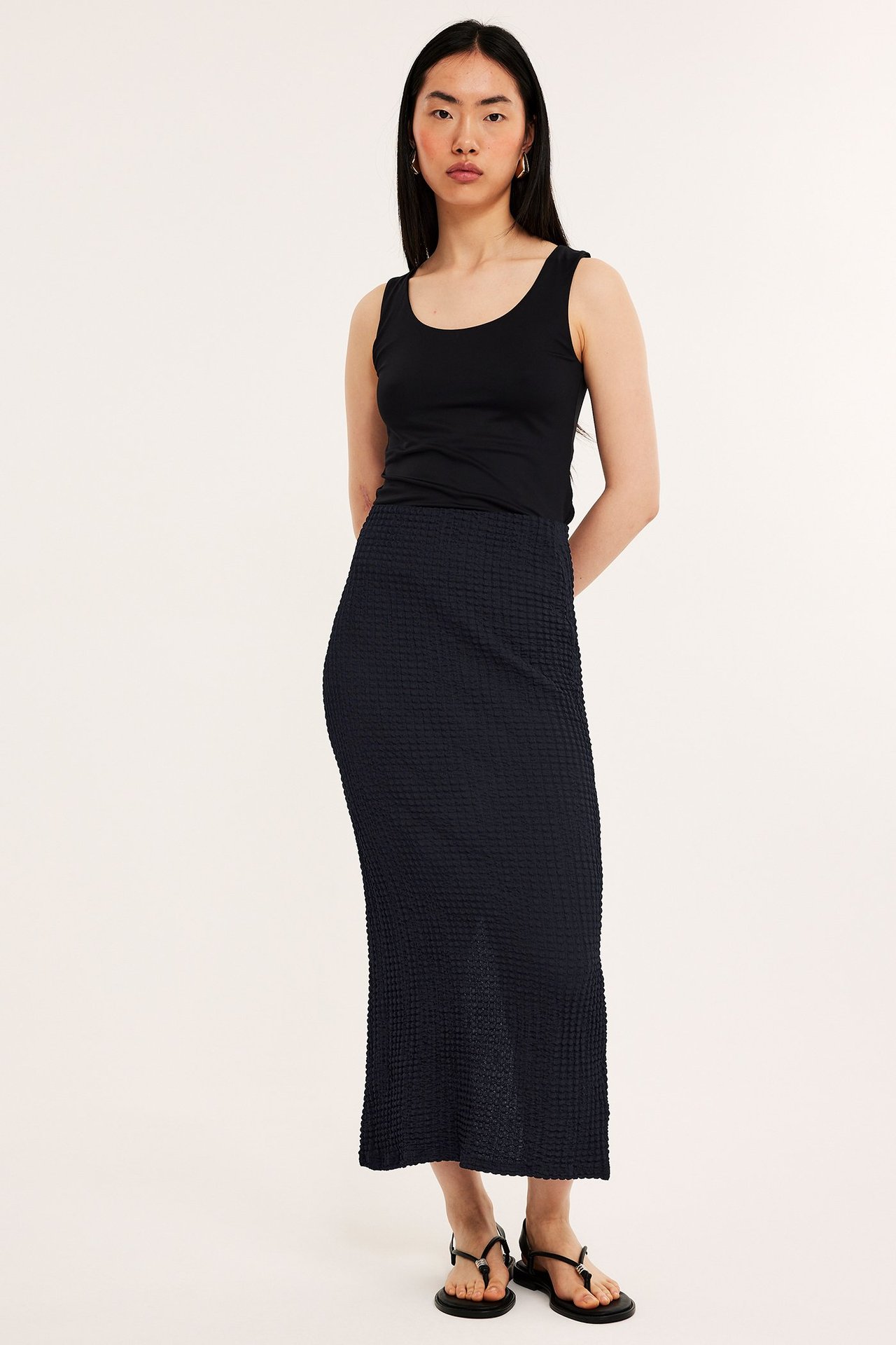 Widok z przodu: kobieta w czarnej strukturalnej długiej spódnicy, czarnym topie i czarnych sandałach.