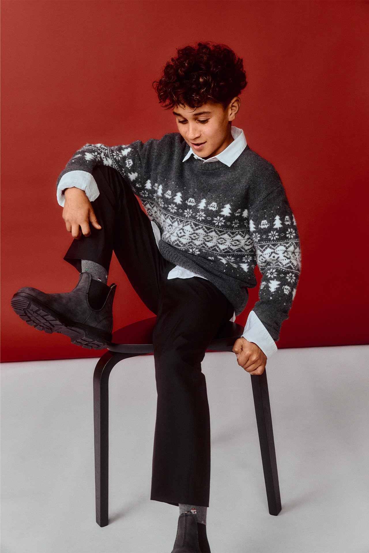 Barn i mörkgrå jacquardstickad jultröja med vita mönster, vit skjorta, svarta byxor och mörkgrå stövlar.
