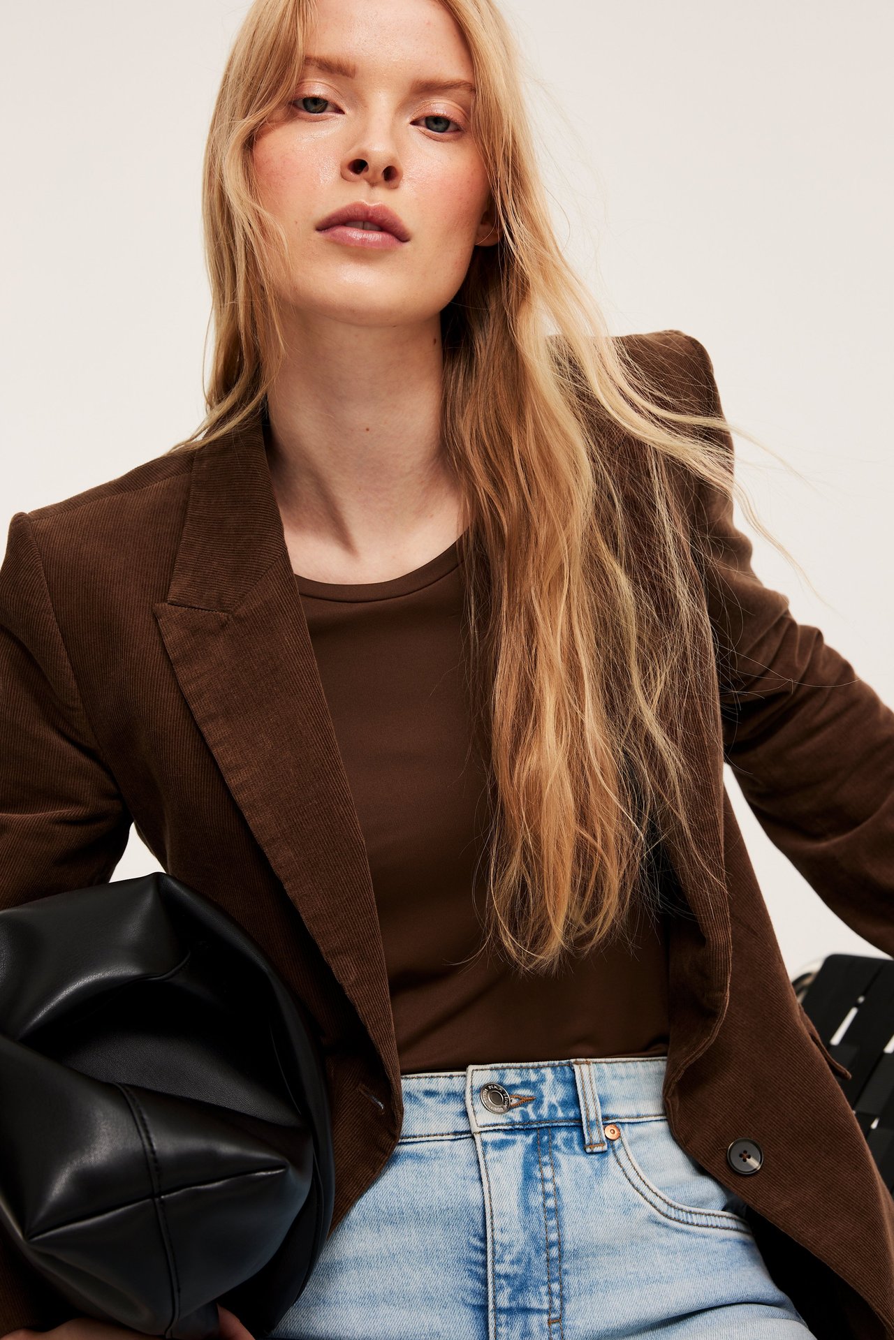 Edestä: Nainen ruskeassa vakosamettibleiserissä, ruskea t-paita, vaaleansiniset farkut, musta laukku.