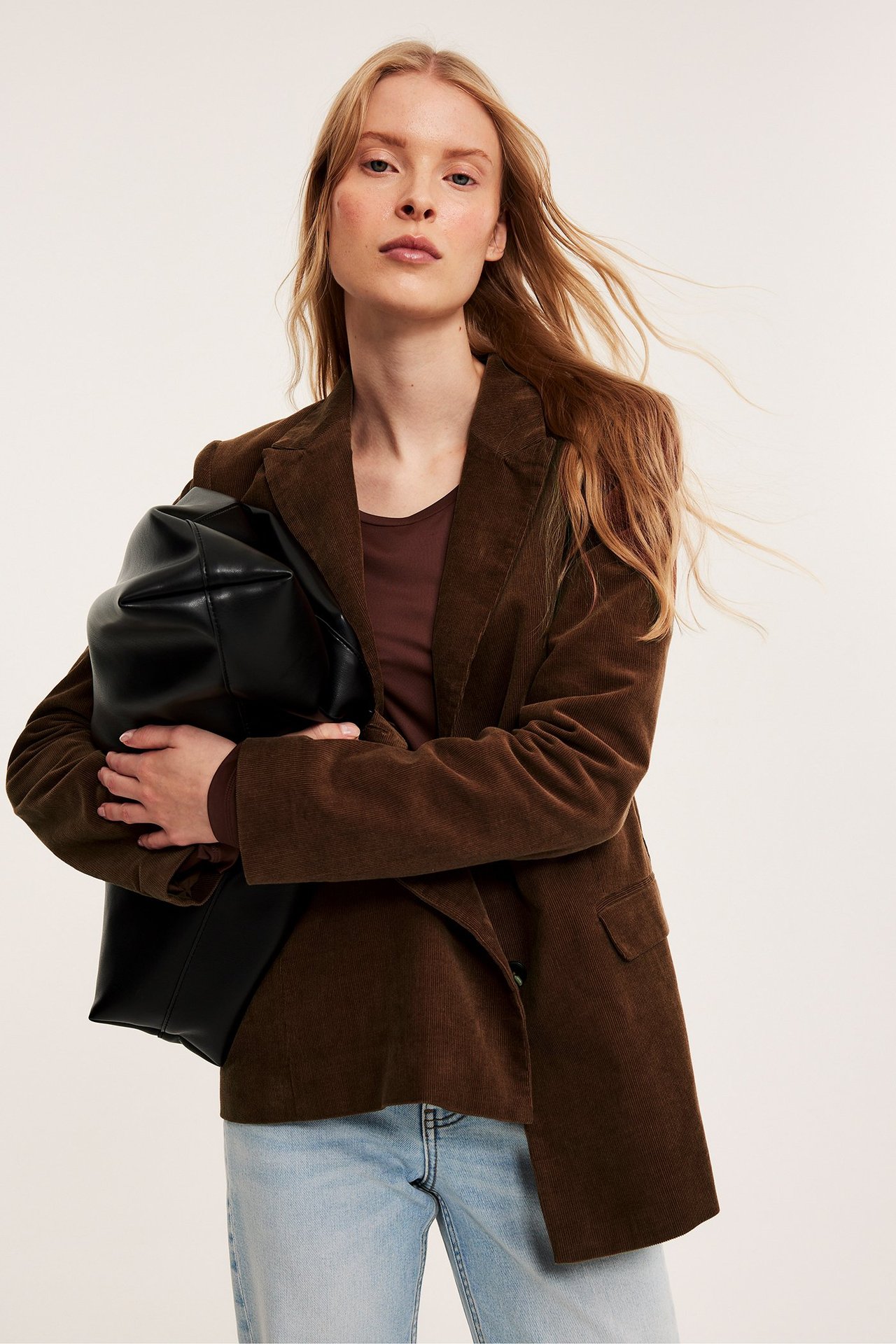 Nainen ruskeassa vakosamettibleiserissä, tummanruskeassa topissa, vaaleansinisissä farkuissa, musta laukku.