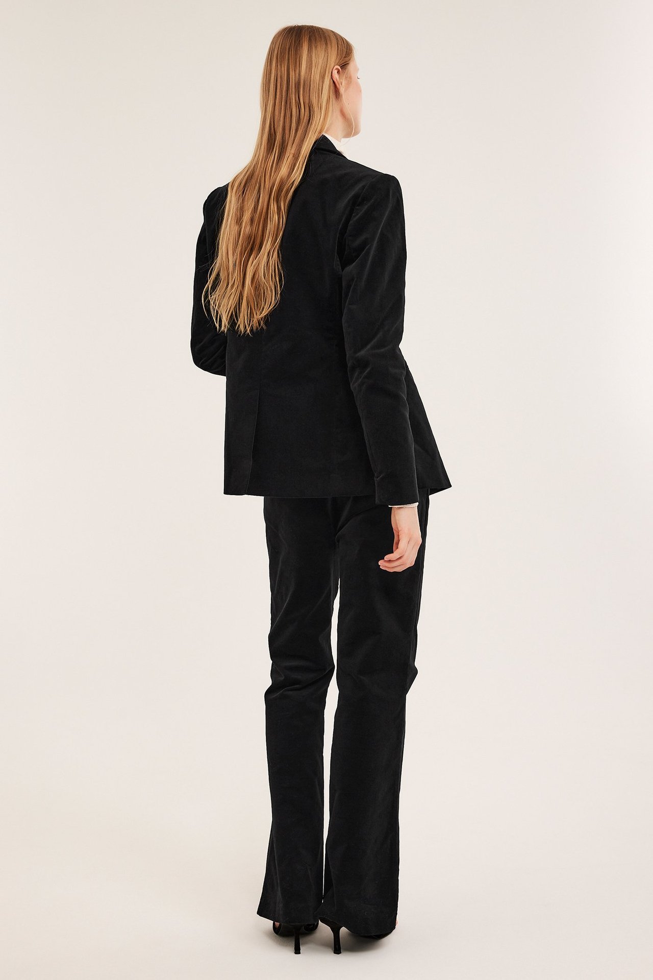 Naisen takakuva mustassa samettibleiserissä ja yhteensopivissa housuissa, mustilla koroilla.