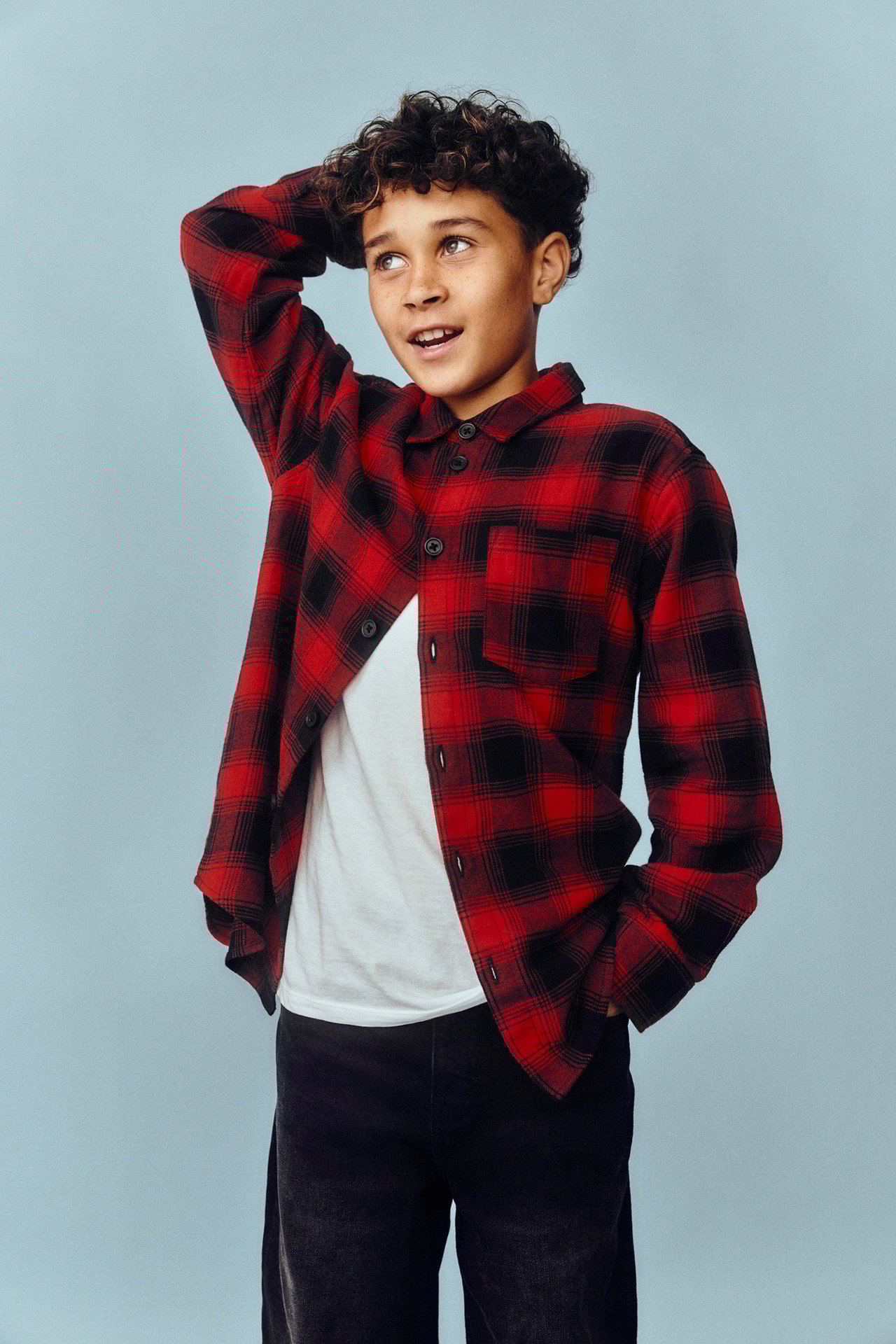 Widok z przodu: dziecko w otwartej czerwonej koszuli flanelowej w kratę, na białym t-shircie i ciemnych spodniach.