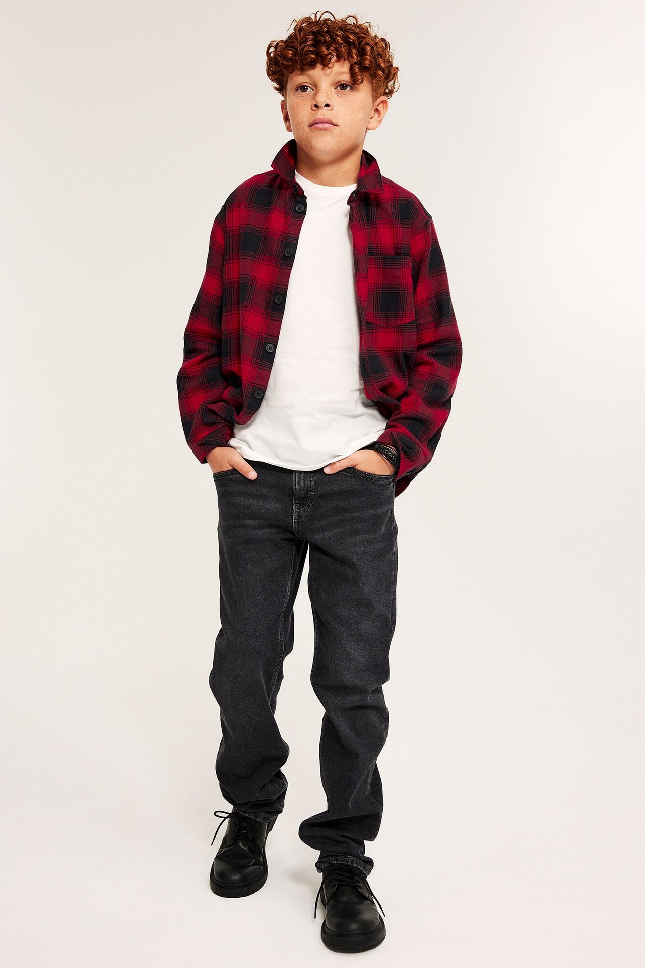 Ung man i svart jeans och svart boots, bär rutig röd flanellskjorta, frontvy.