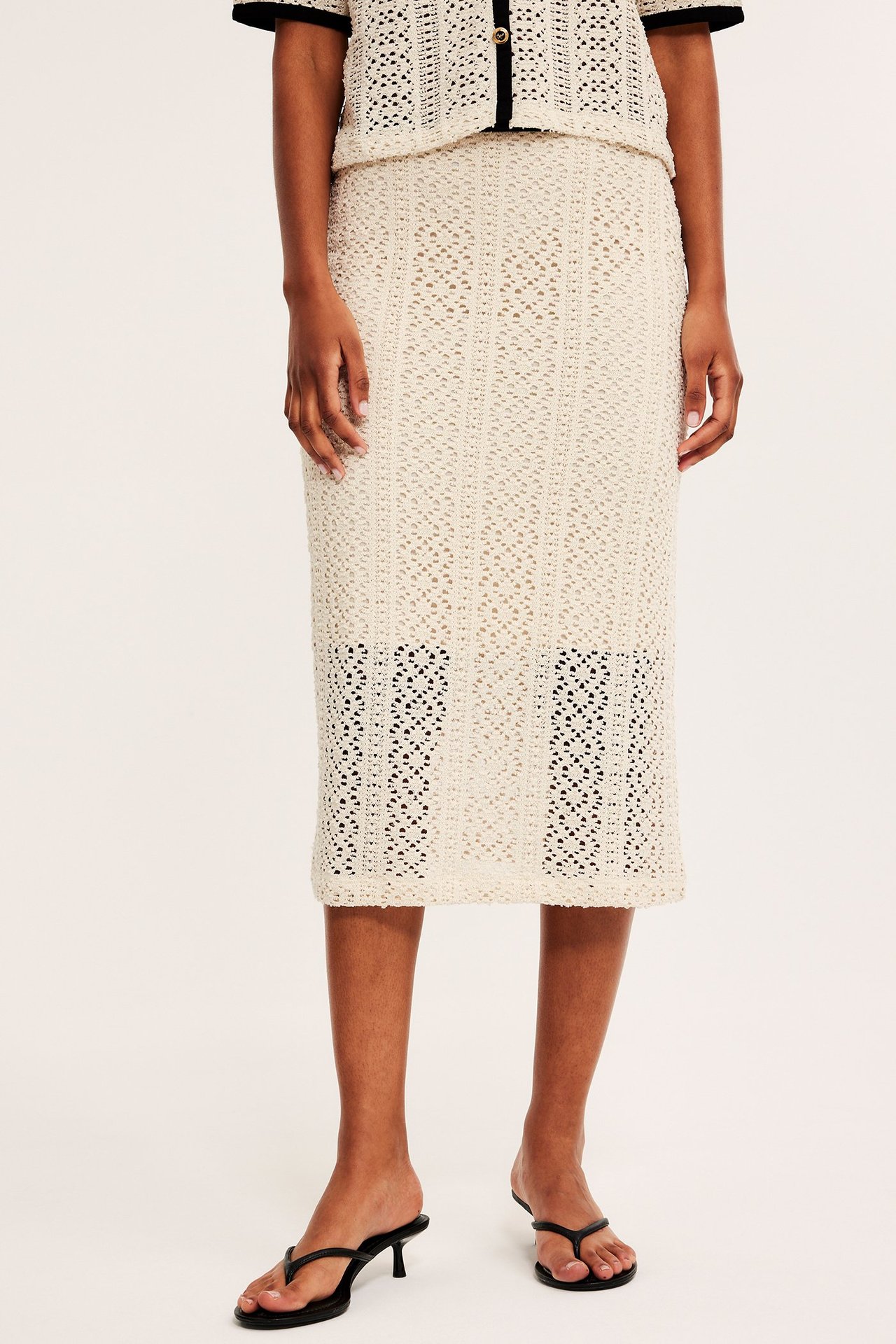 Forfra, kvinne i offwhite heklet midiskjørt, matchende topp og svarte høye sandaler.