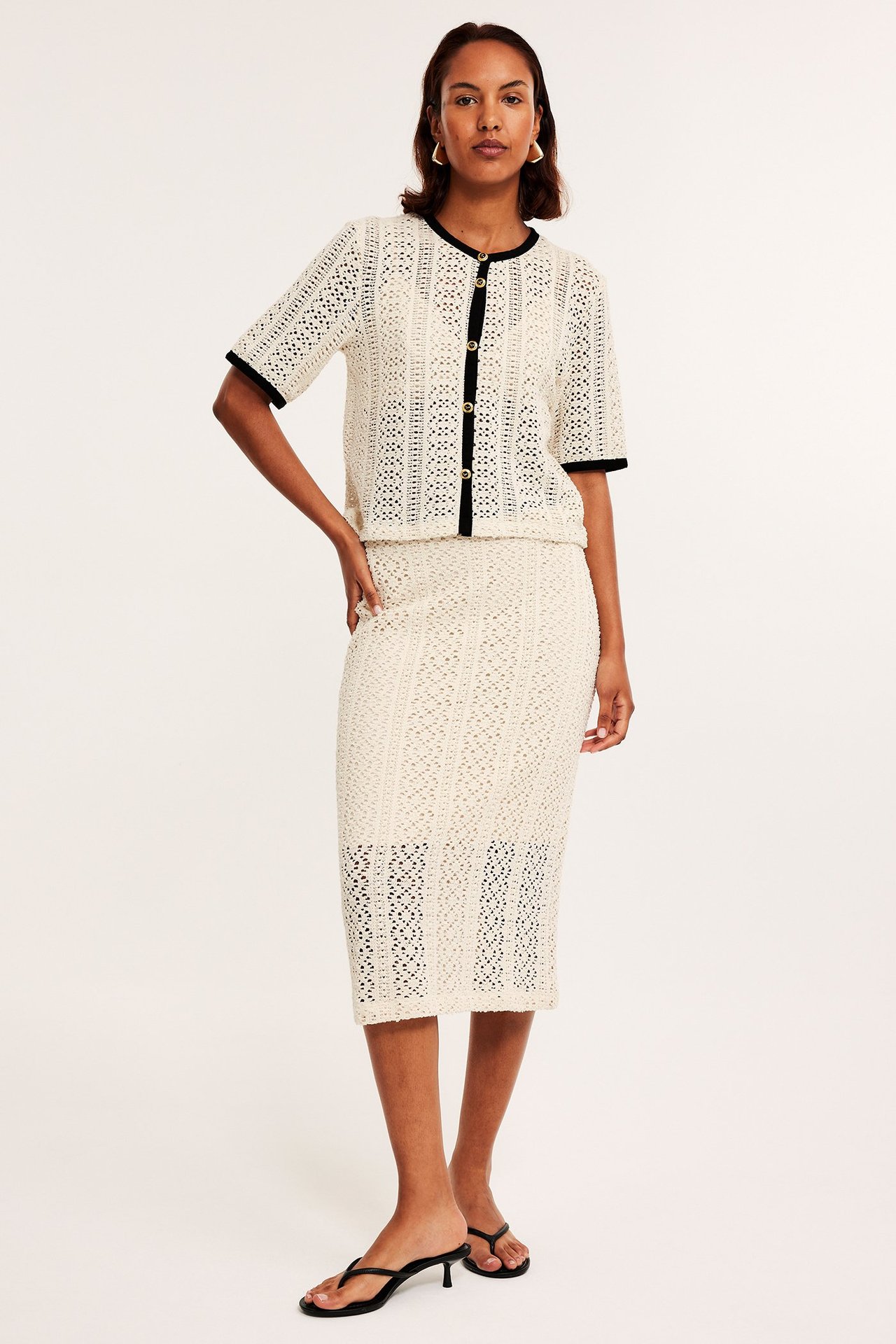 Widok z przodu: Kobieta w kremowej szydełkowej spódnicy midi, pasującym topie i czarnych sandałach na obcasie.