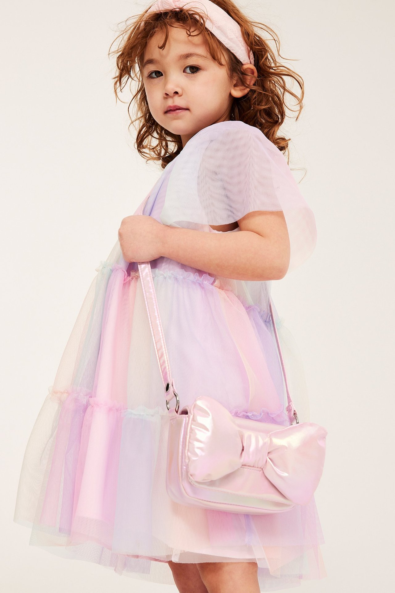 Dziecko nosi jasnoróżową opalizującą torebkę z kokardą, tęczową sukienkę tiulową i różową opaskę.