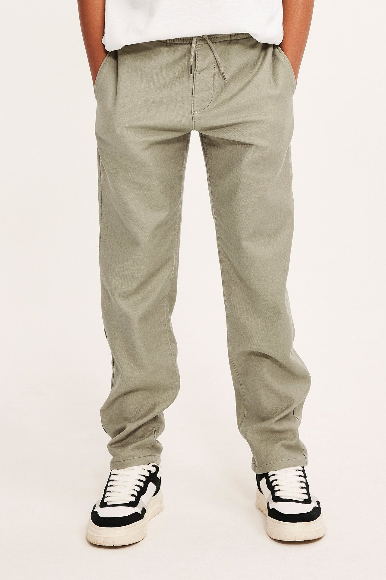 Lapsi yllään khakinvihreät kiristysnyöri-chinosit, valkoinen t-paita ja mustavalkoiset tennarit, edestäpäin.