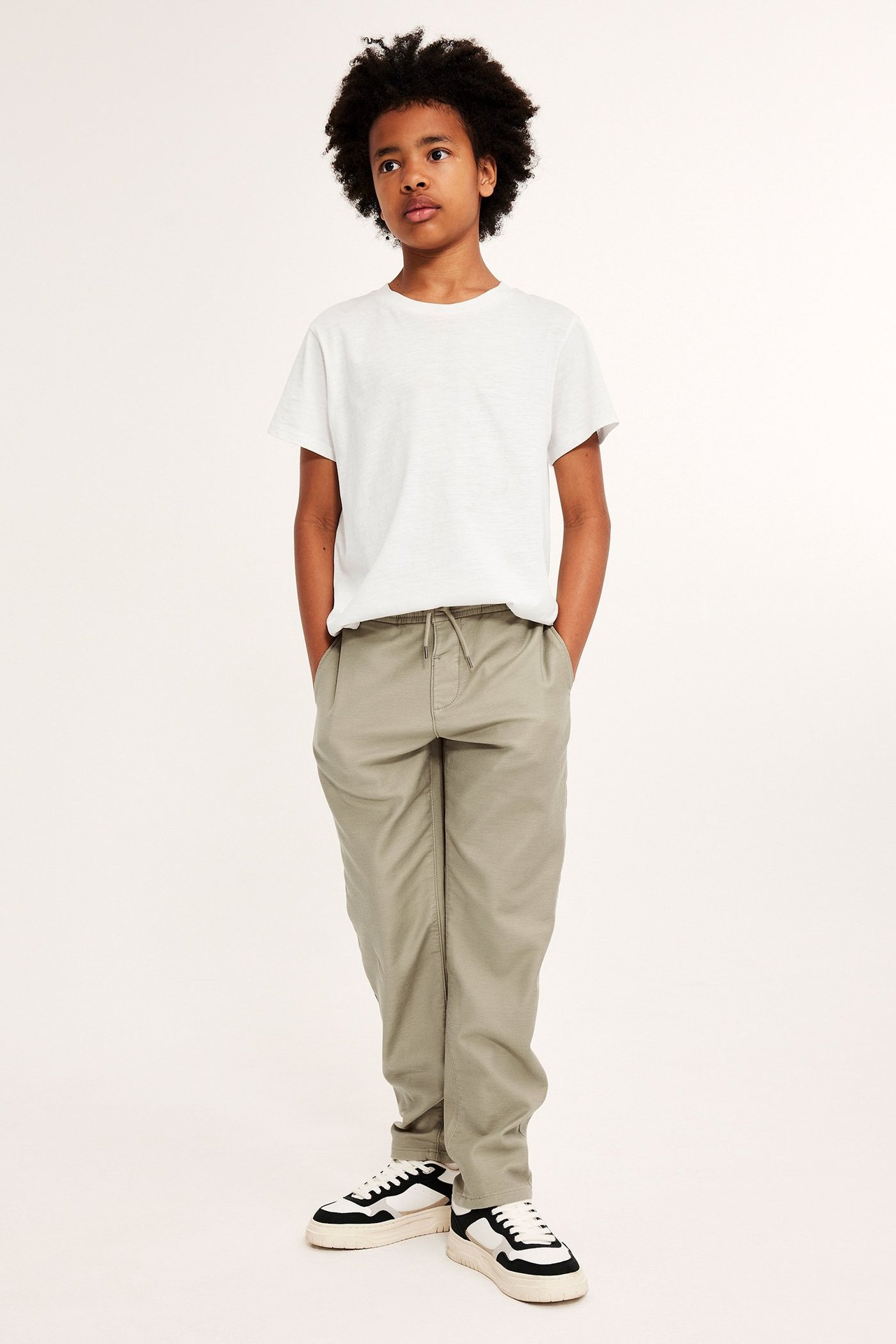 Widok z przodu: dziecko w spodniach chino w kolorze khaki z troczkiem, białej koszulce i czarno-białych sneakersach.