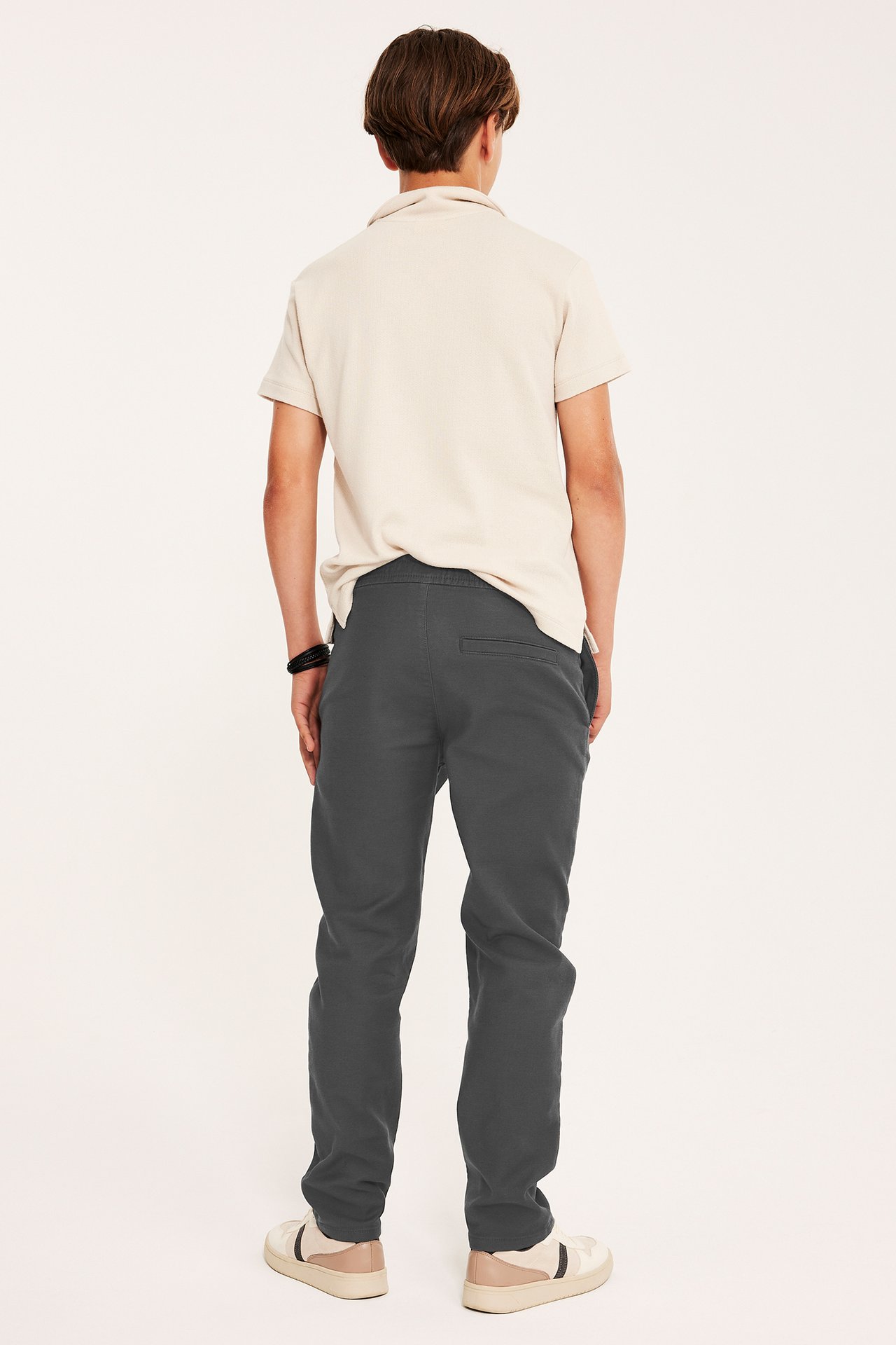 Bakifrån: ett barn i svarta stretchchinos med resår i midjan, en beige pikétröja och beiga sneakers.