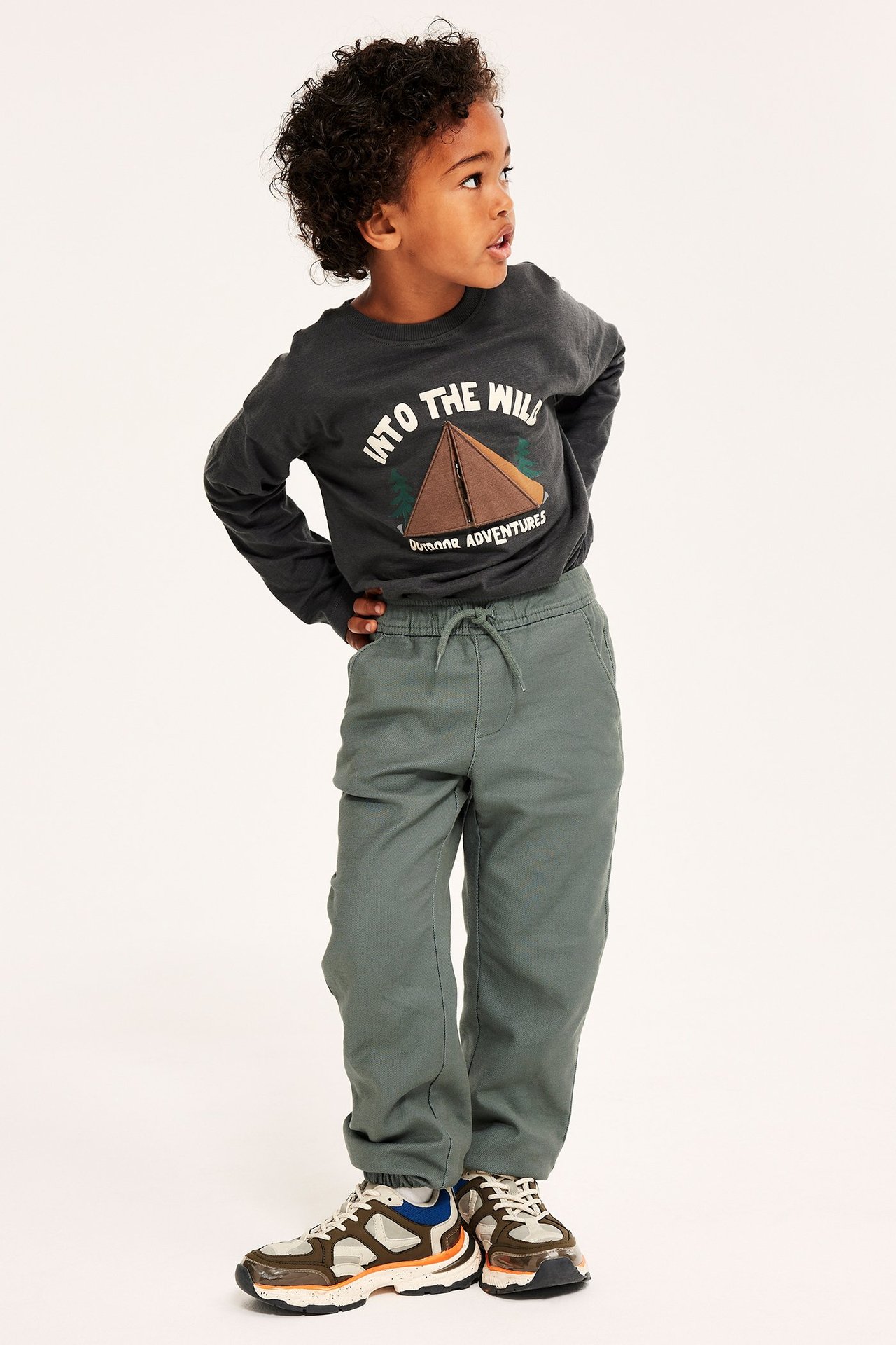 Edestäpäin lapsi yllään tummanvihreät kiristysnyörilliset chinot, tummanharmaa pitkähihainen paita ja moniväriset lenkkarit.