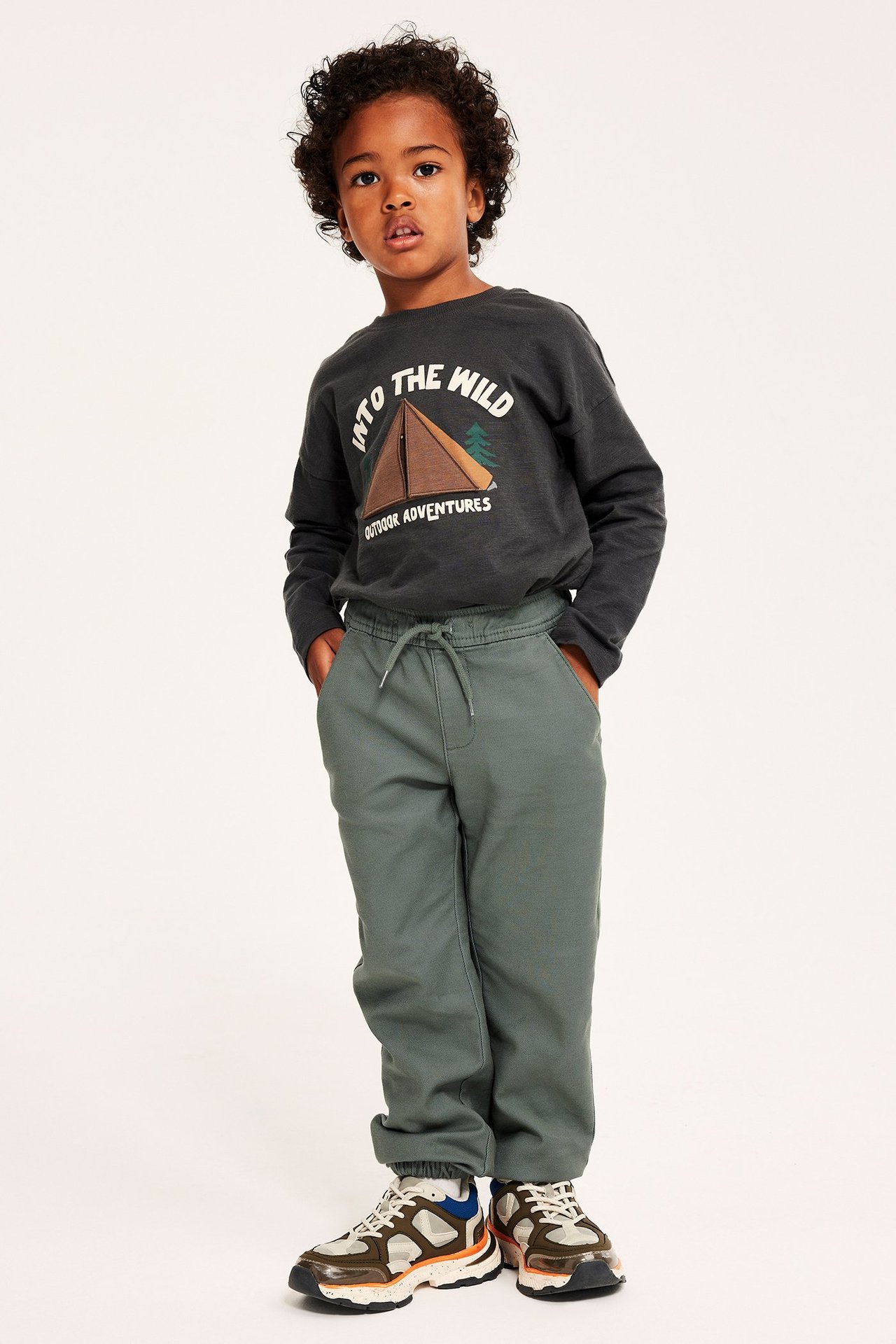 Edestä kuvattu lapsi tummanvihreissä joustavissa chinoissa kiristysnyörillä, tummanharmaassa pitkähihaisessa paidassa ja lenkkareissa.