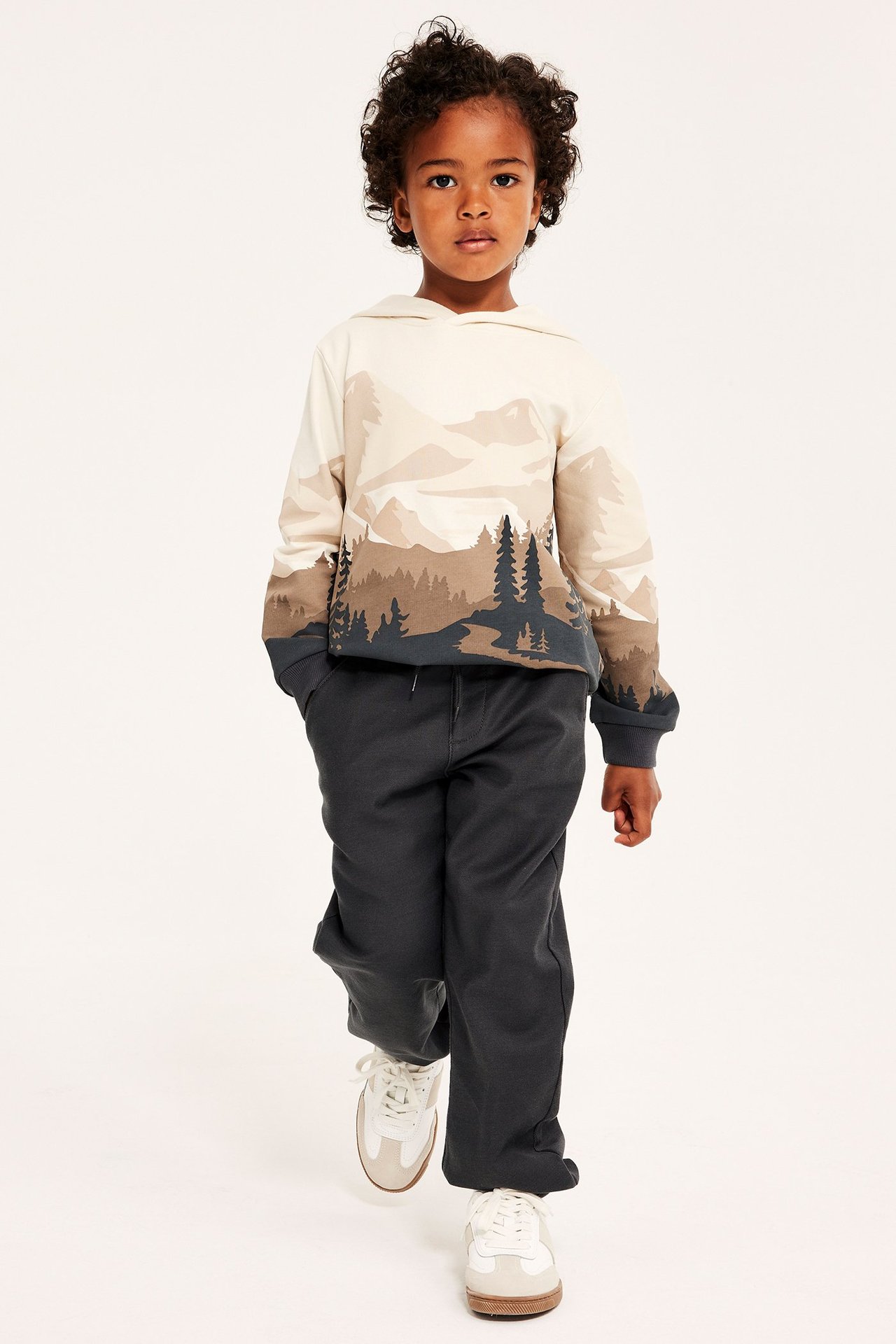 Forfra, et barn i svarte myke chinosbukser med snøring, en hettegenser med fjellmønster og hvite joggesko.