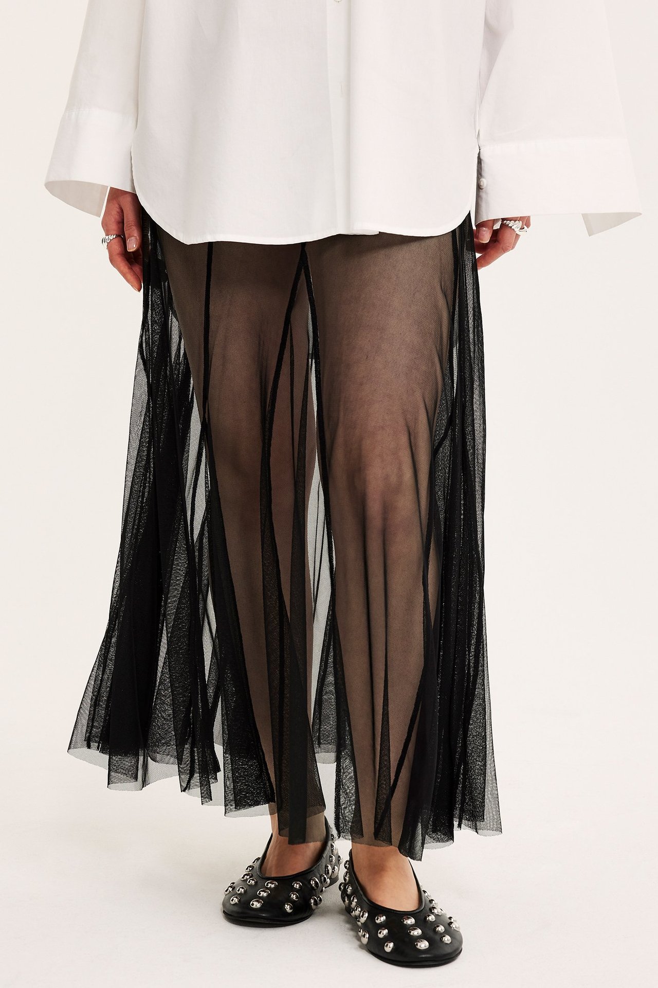 Naisen etunäkymä mustassa pitkässä paneelihameessa, valkoisen paidan ja niittikoristeisten kenkien kanssa.