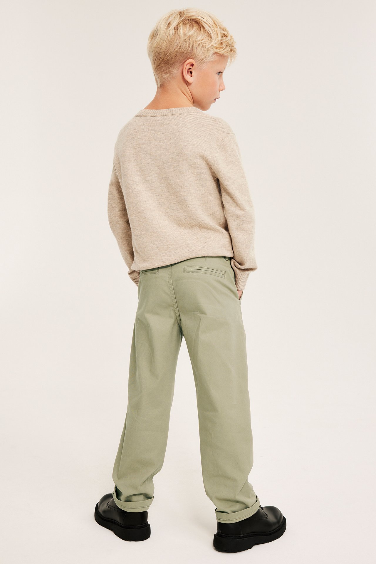 Bakfra, et barn i løse khakigrønne chinos, en beige genser og svarte kraftige sko.