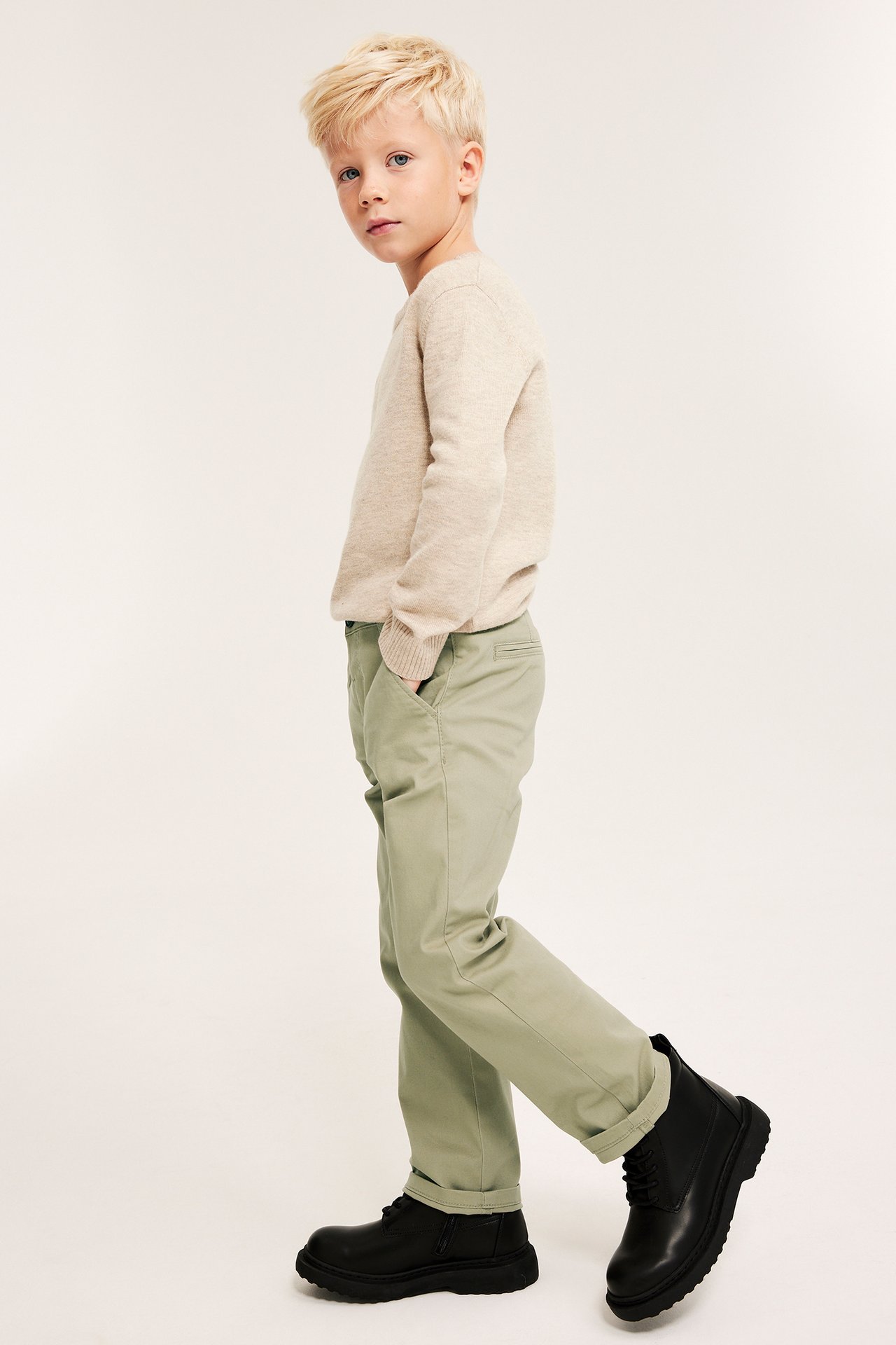 Ung mann i beige genser og lysegrønne chinos, svarte boots, sidevisning.
