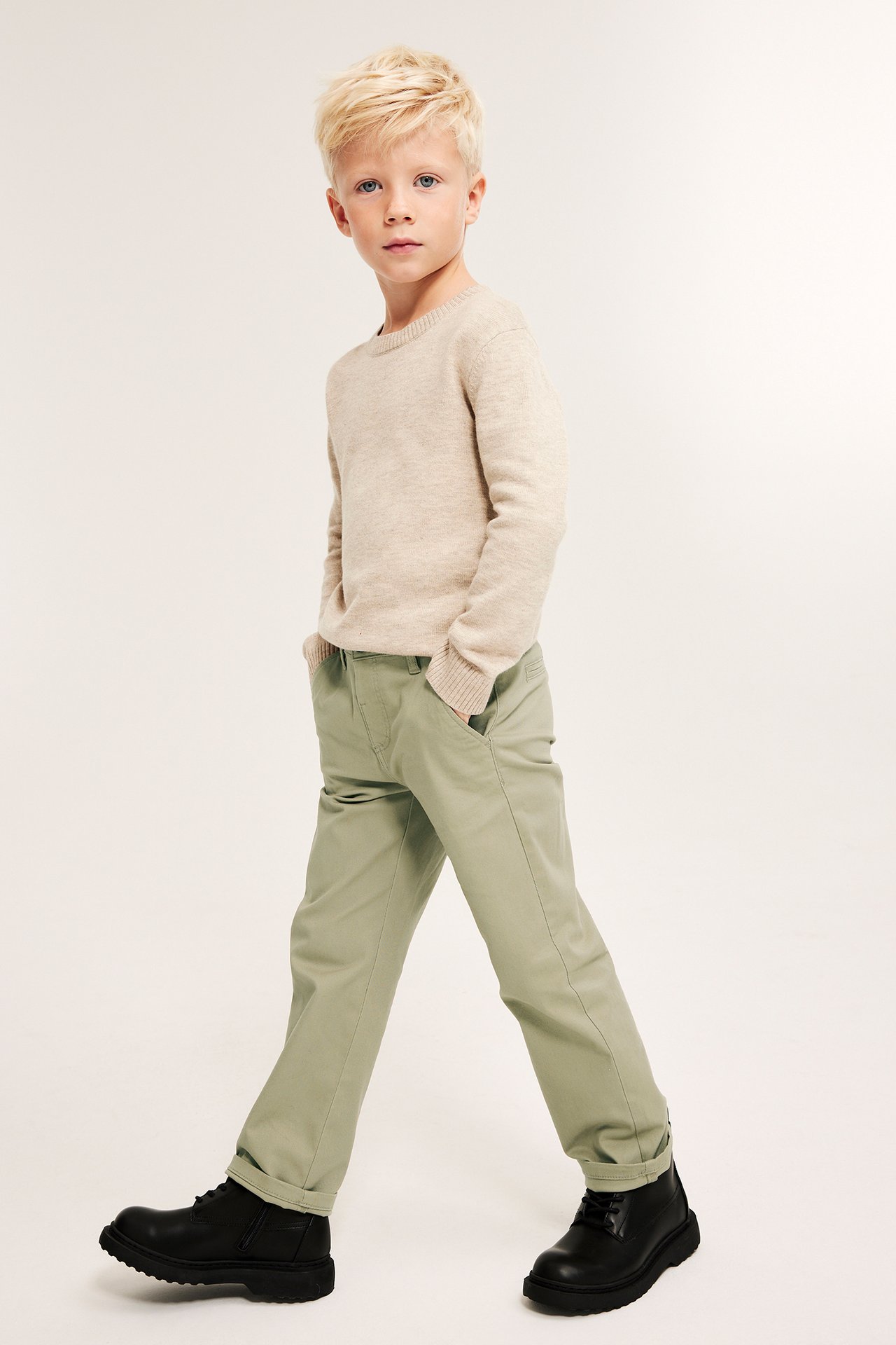 Frontvisning av barn i lysegrønne stretchchinos, en beige genser og svarte boots.