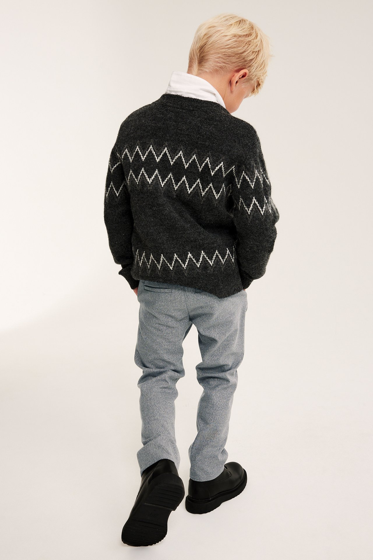Barn från ryggen i ljusgrå melerade kostymbyxor, mörkgrå zigzagsweater, vit topp och svarta skor.