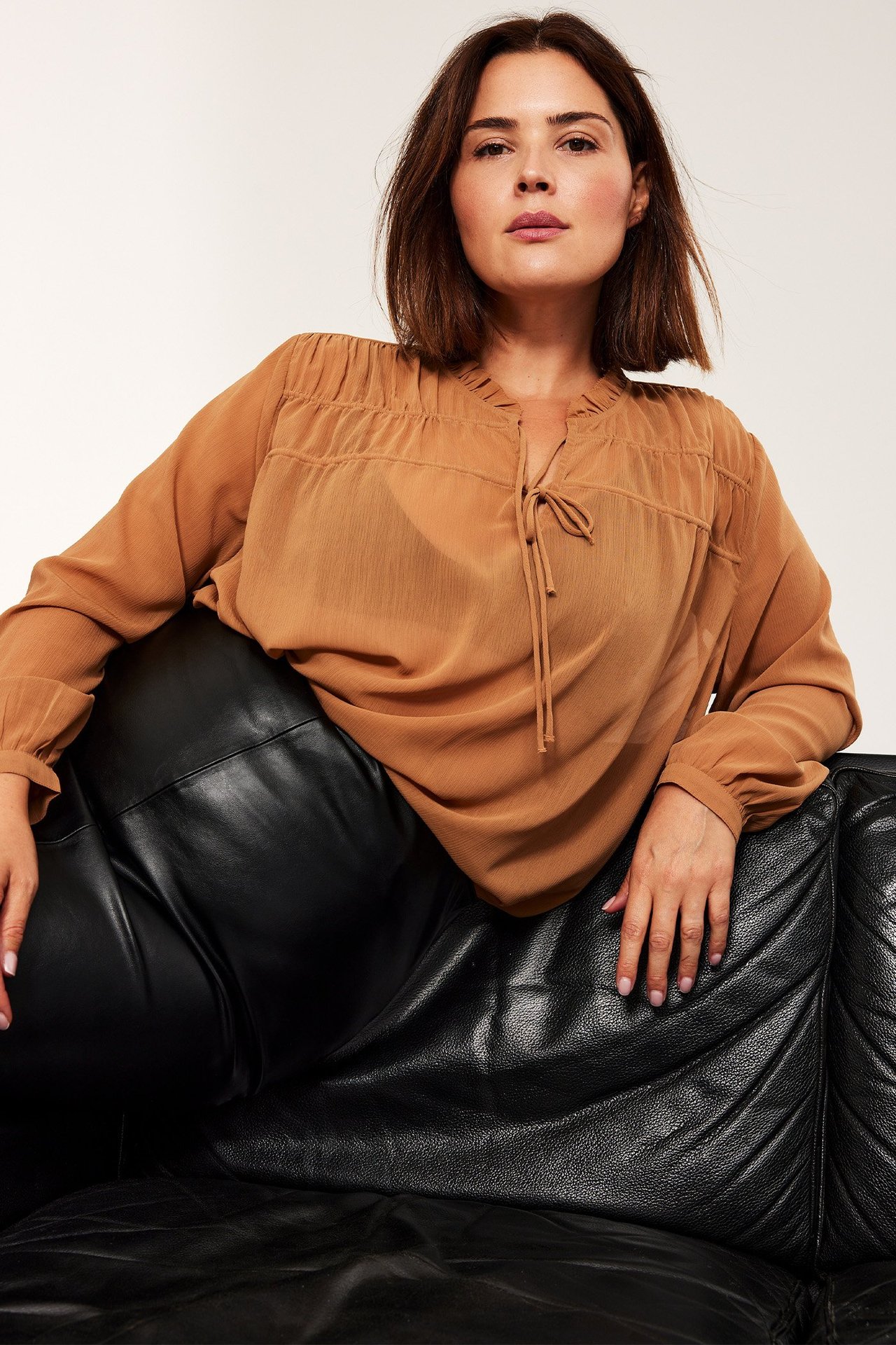 Frontbilde av kvinne i gjennomsiktig beige langermet polyesterbluse med knyting og sorte bukser.