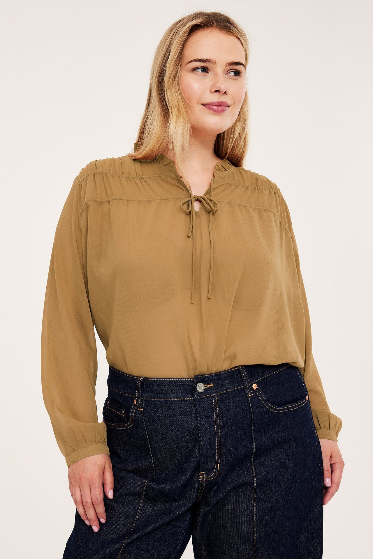 Edestä näkymä naisesta beigessä pitkähihaisessa puserossa nyörityksellä ja tummansinisissä farkuissa.