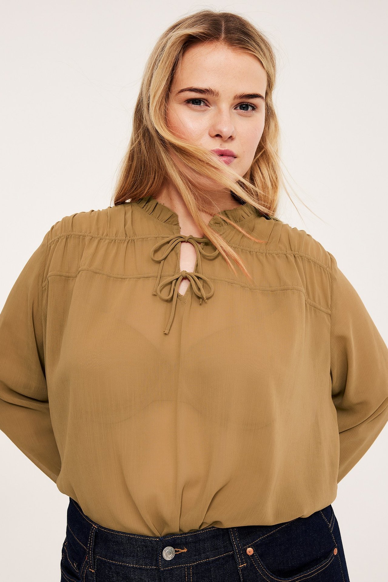 Widok z przodu, kobieta w luźnej beżowej prześwitującej bluzce ze ściągaczem, na ciemnych dżinsach.