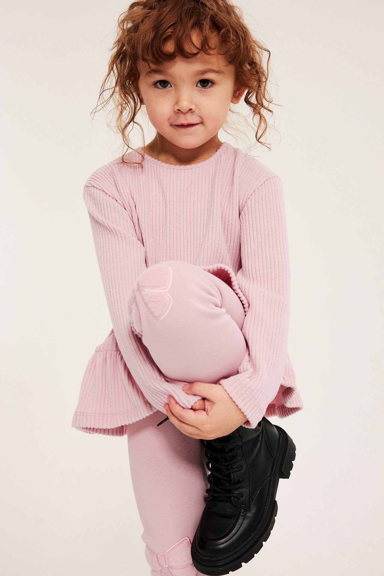 Front view av ett barn i ljusrosa värmande leggings med rosetter, rosa ribbad topp och svarta kängor.
