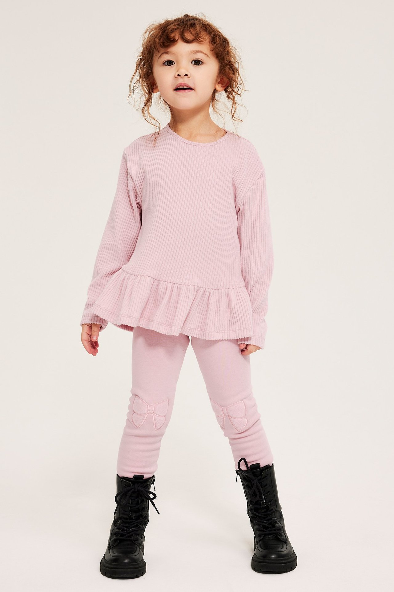 Dziecko nosi jasnoróżowe ocieplane legginsy z kokardami, różowy prążkowany top z falbaną i czarne buty.