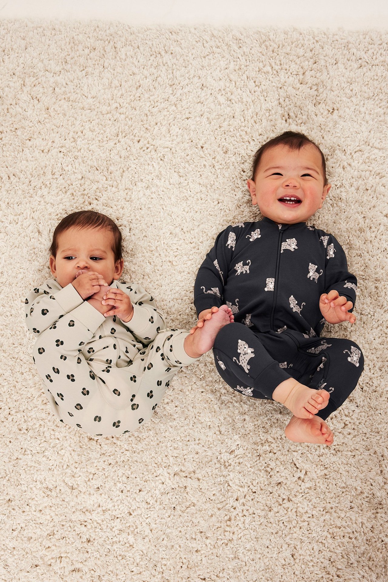 Toppvy av två bebisar i leopardmönstrade pyjamasar: en ljusbeige, en mörkgrå, båda med tvåvägsdragkedja.