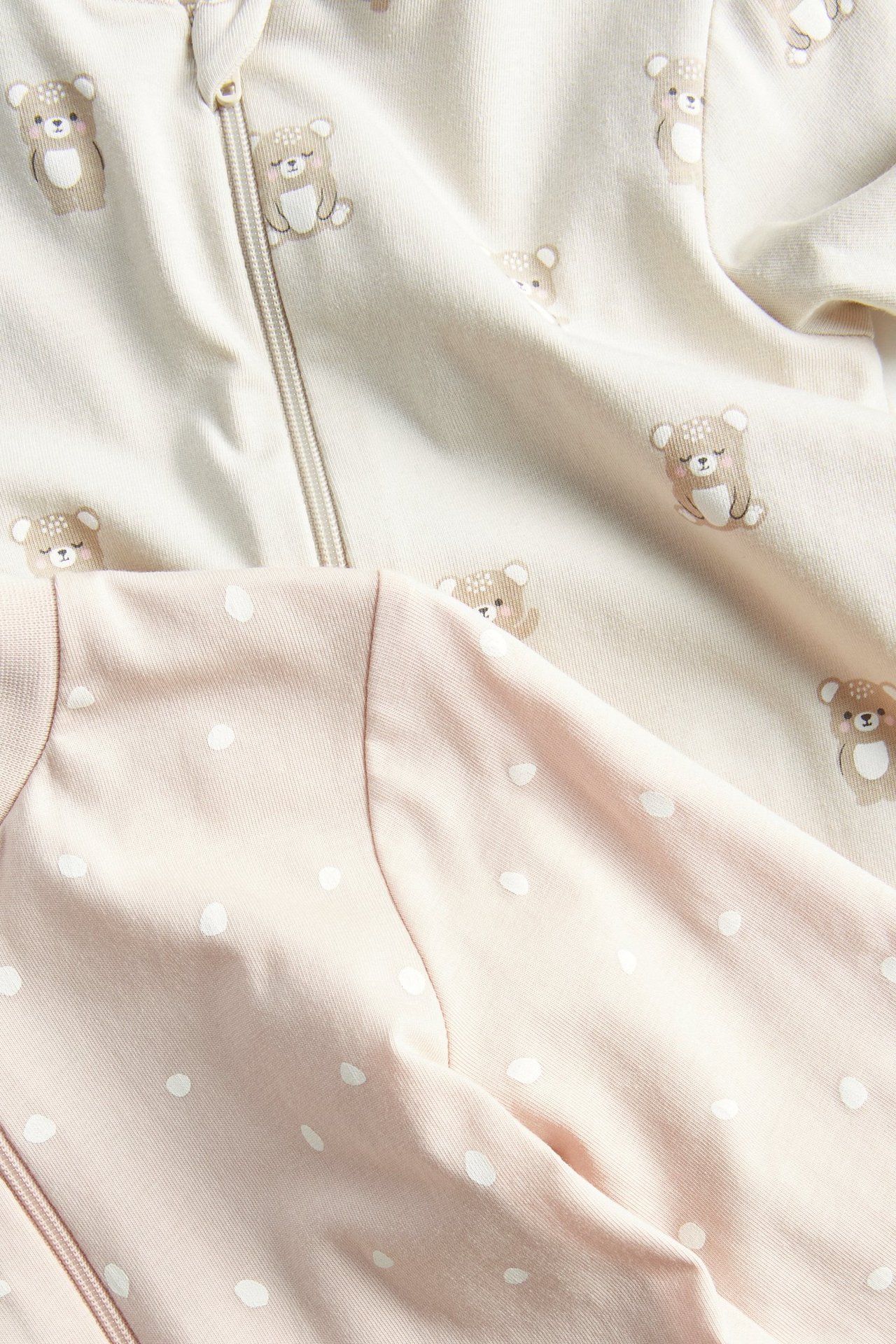 Zbliżenie dwóch pajacyków niemowlęcych: beżowego w misie z zamkiem i różowego w białe kropki.