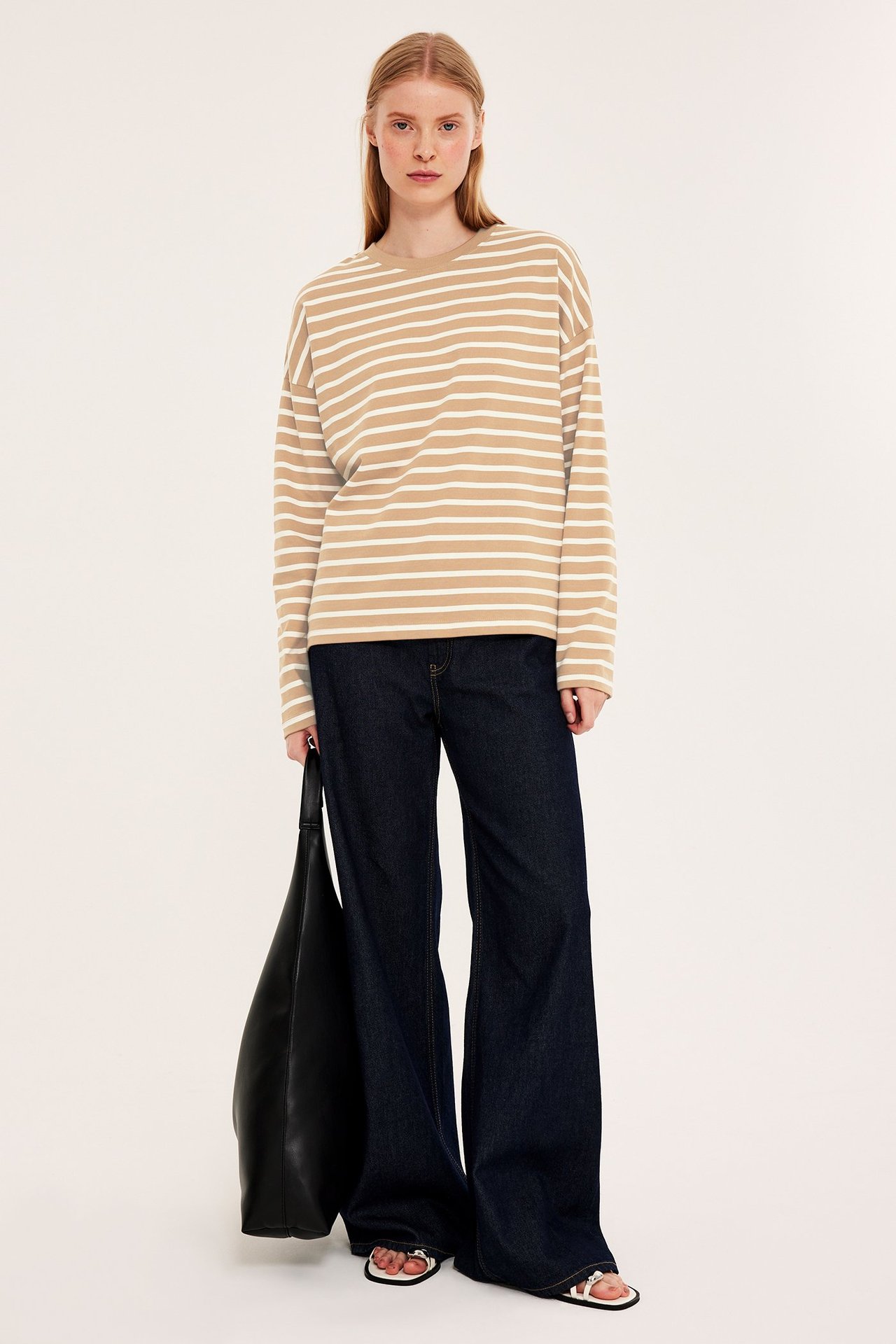 Forfra: Kvinne i løs beige og hvitstripet bomullstopp, mørkeblå jeans, hvite sandaler, holder svart veske.