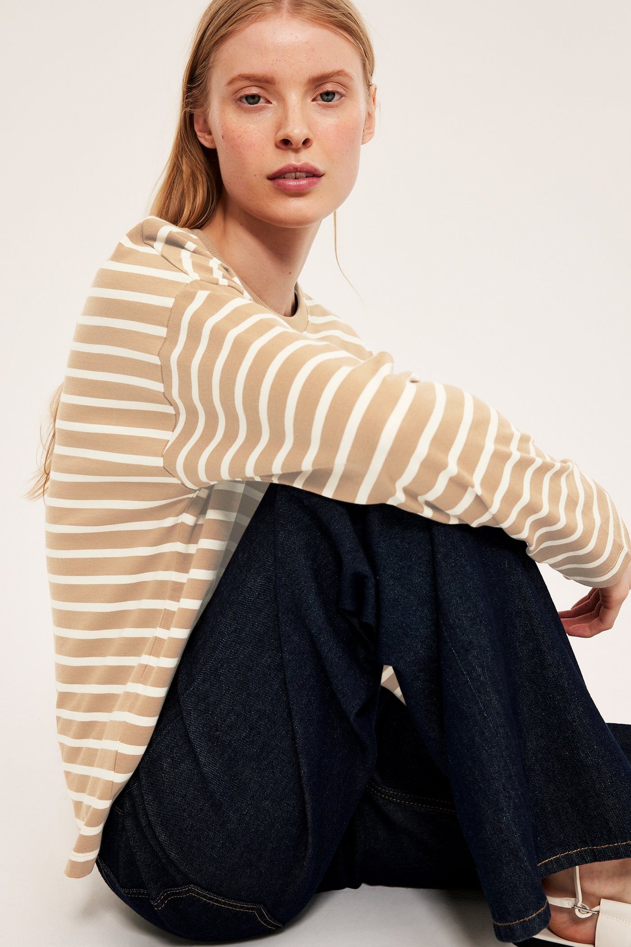 Widok z przodu: kobieta w beżowo-białej bluzce w paski z długim rękawem i ciemnoniebieskich dżinsach.