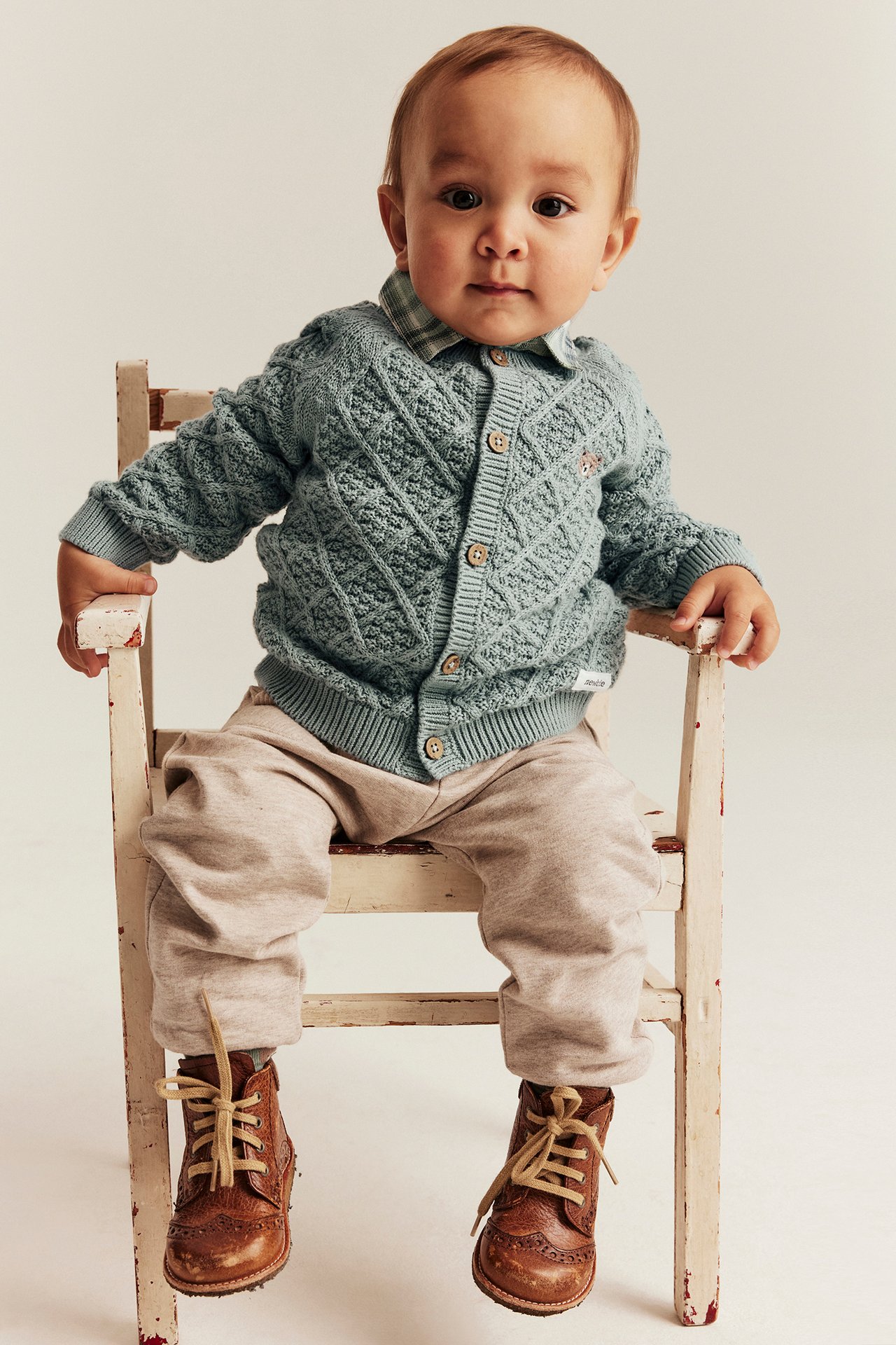 Forfra: Baby i lysegrønn kabelstrikket jakke med bamseapplikasjon, over rutete skjorte, beige bukser og brune sko.