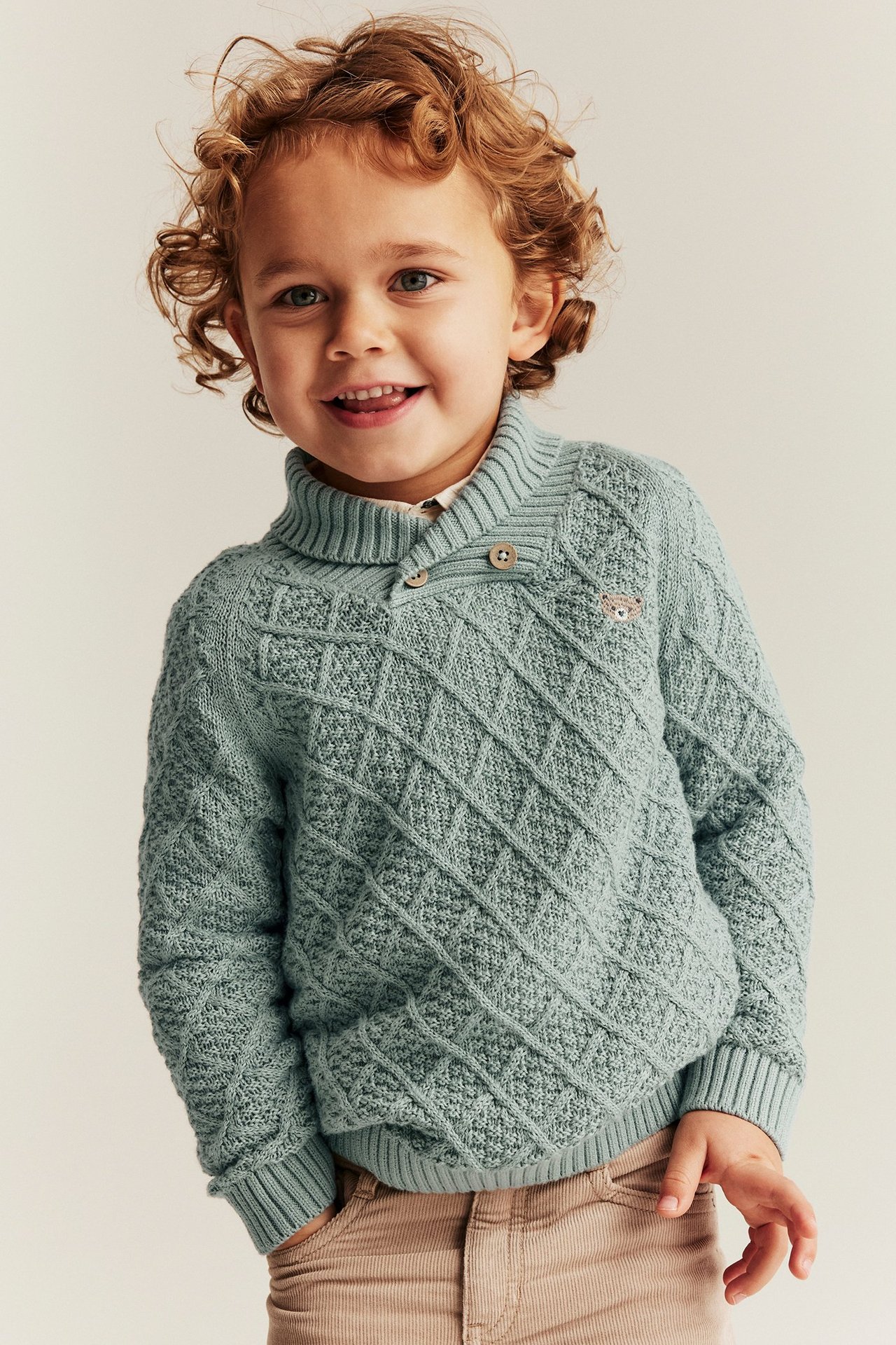 Forfra, et barn i lysegrønn strikkegenser med knappekrage, teddybroderi og beige bukser.