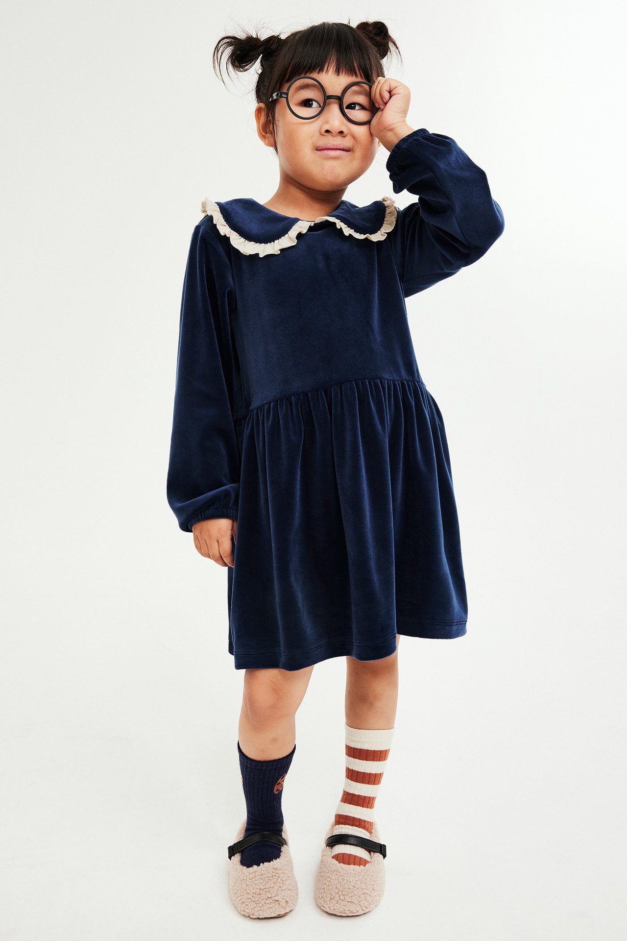 Frontbilde av et barn i mørk blå velourkjole med kremfarget rysjekrage, ulike sokker og lodne tøfler.
