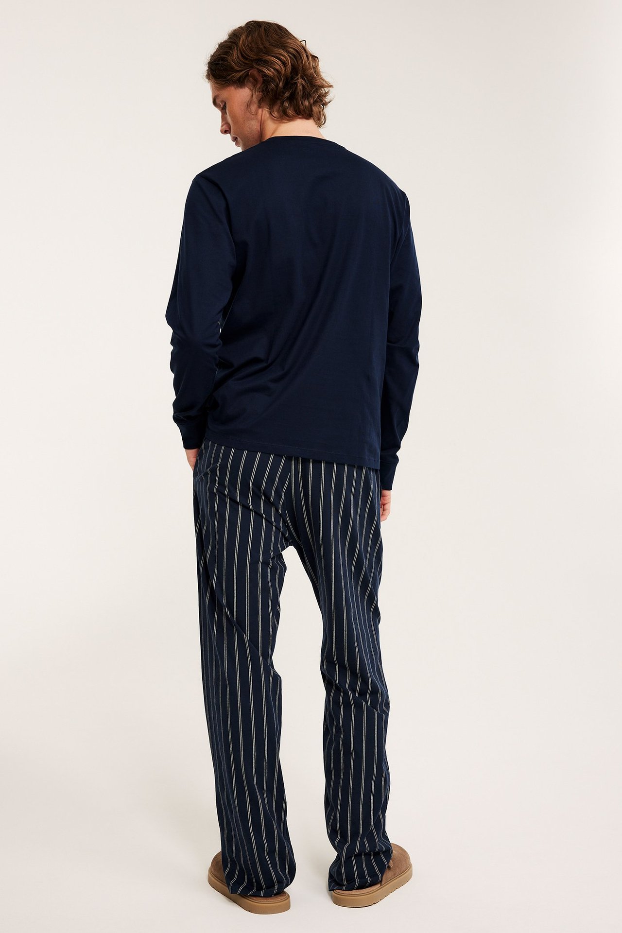 Takakuva: Mies tummansinisissä raidallisissa pyjamahousuissa, tummansinisessä paidassa ja ruskeissa tossuissa.