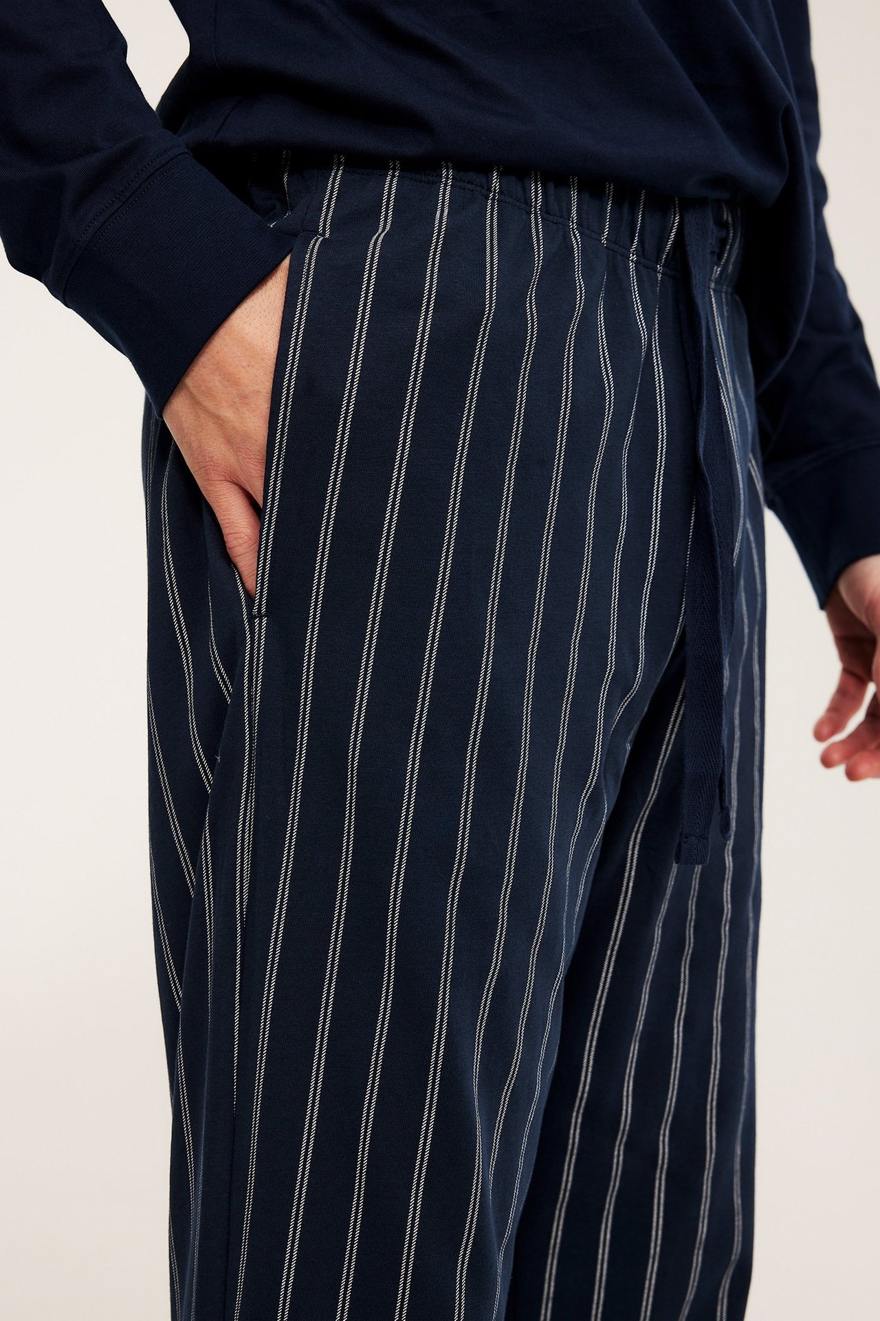 Närbild på man i mörkblå randiga bomullspyjamasbyxor med dragsko, hand i ficka, och mörkblå topp.