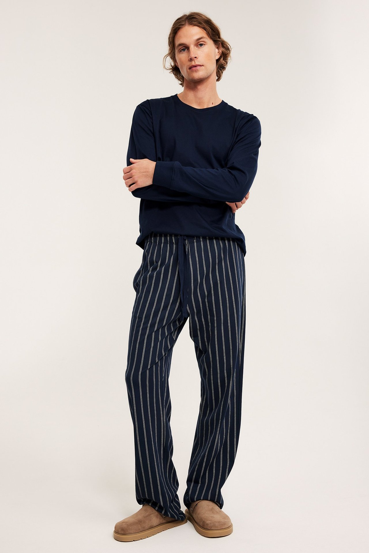Widok z przodu: mężczyzna w granatowych, pasiastych spodniach piżamowych, granatowym longsleeve'ie i brązowych kapciach.