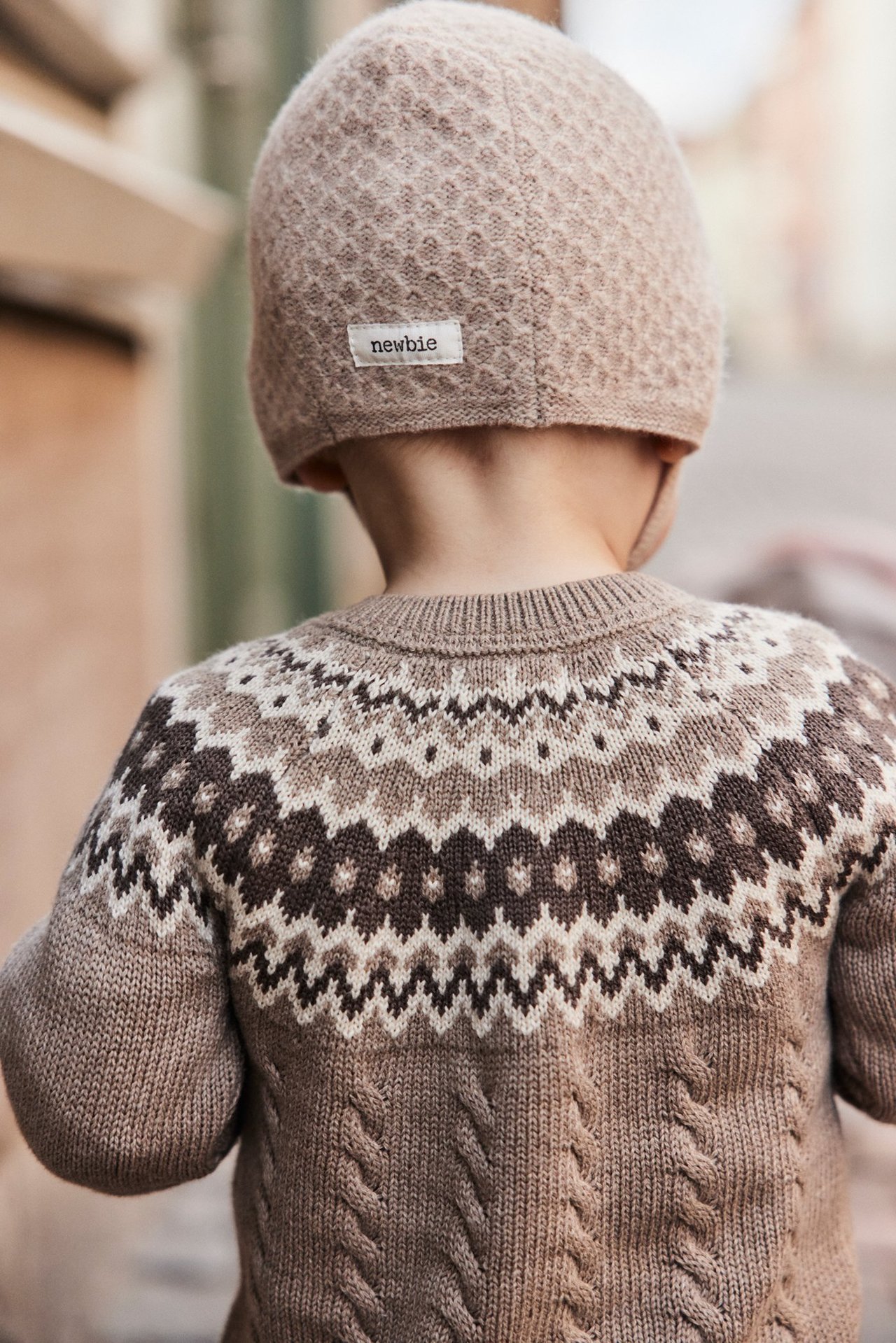Bakifrån: Baby i beige mönsterstickad hjälmmössa och beige tröja med fair isle-mönster.
