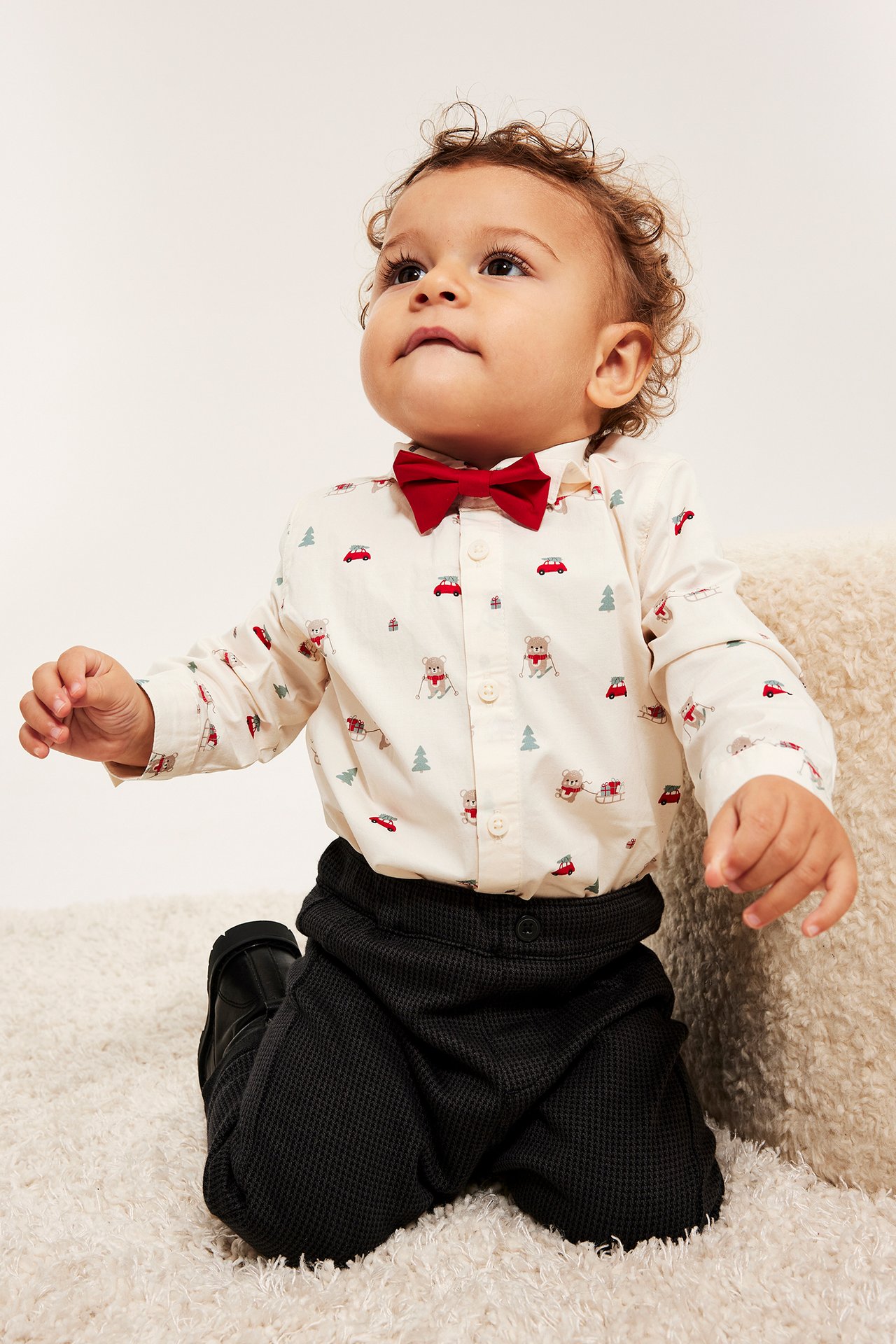 Baby i offwhite julebluse med rød sløyfe, mønster av hunder og biler, svarte bukser.