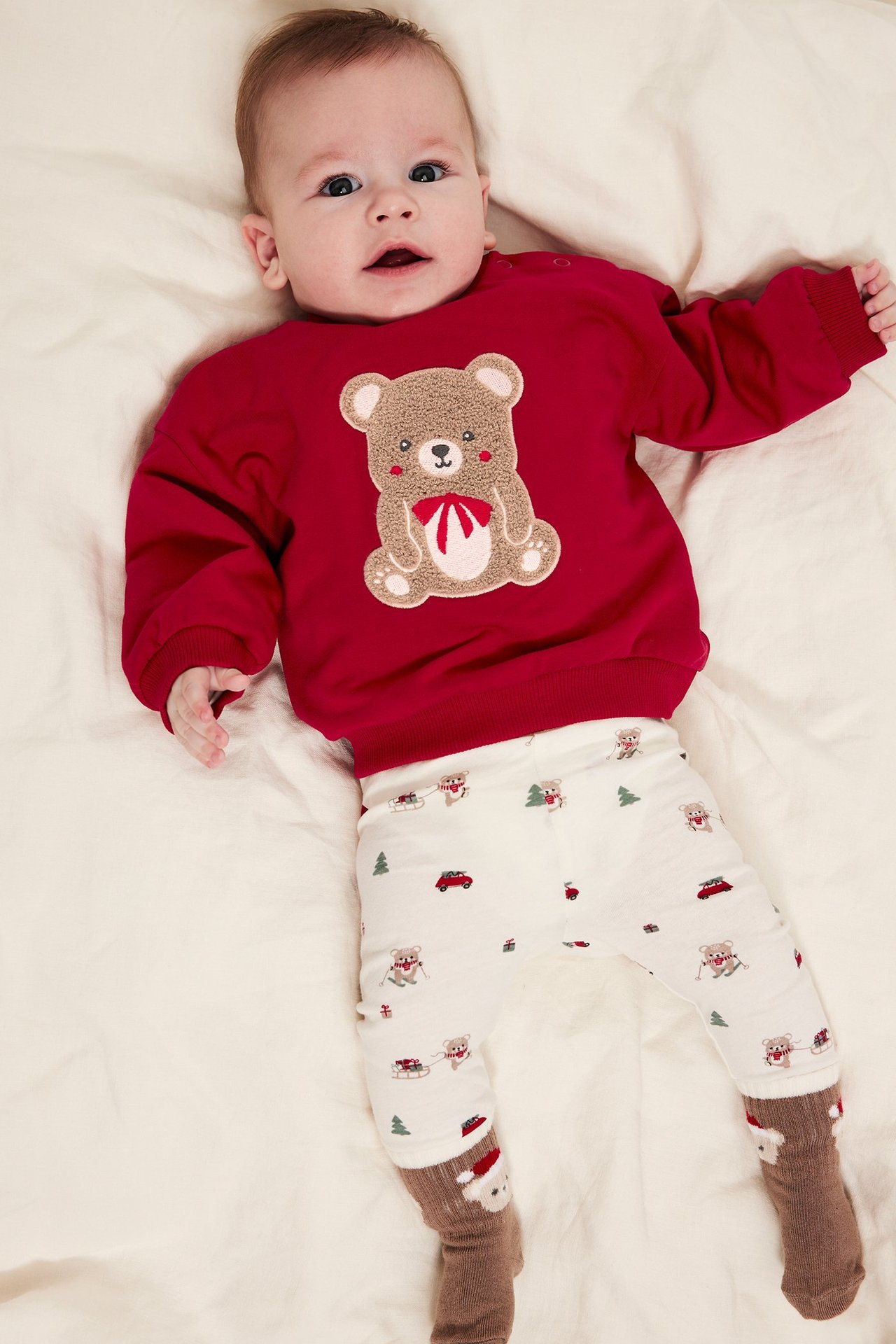 Baby i rød sweatshirt med julmotiv og trykk foran, ligger på seng, frontview.