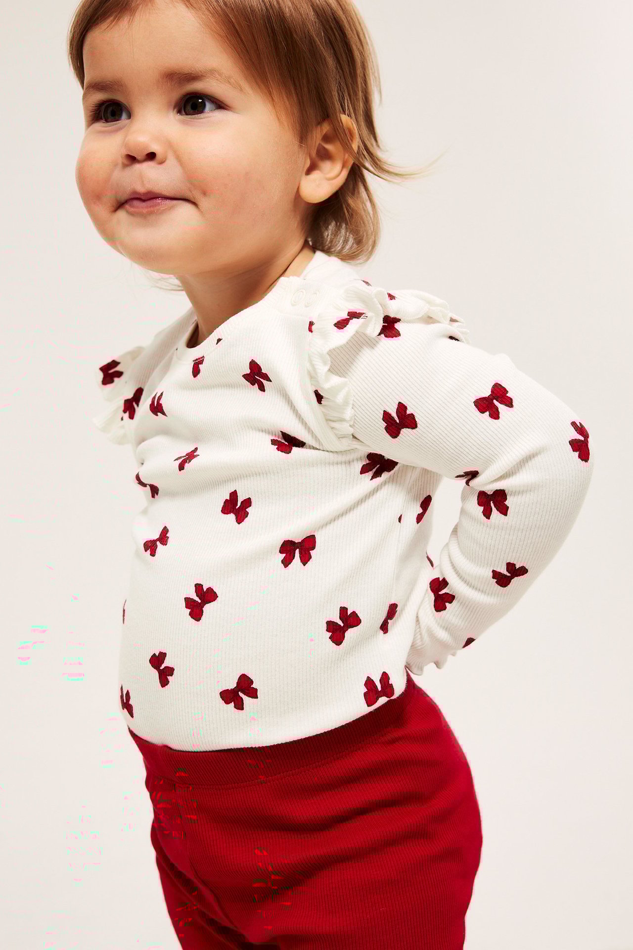 Dziecko w białym prążkowanym body z czerwonymi kokardami, bok widok, zatrzaski.