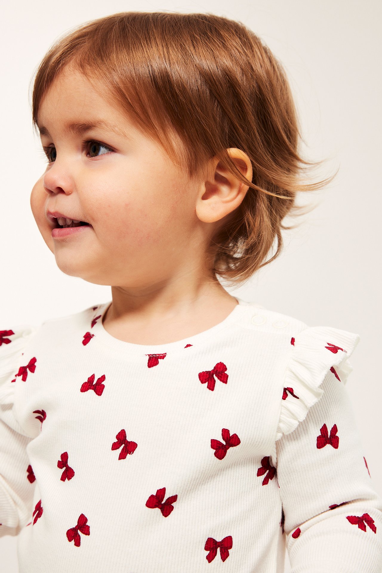 Dziewczynka w kremowej, prążkowanej body z czerwonymi kokardkami, zatrzaskami na ramionach i kroku.